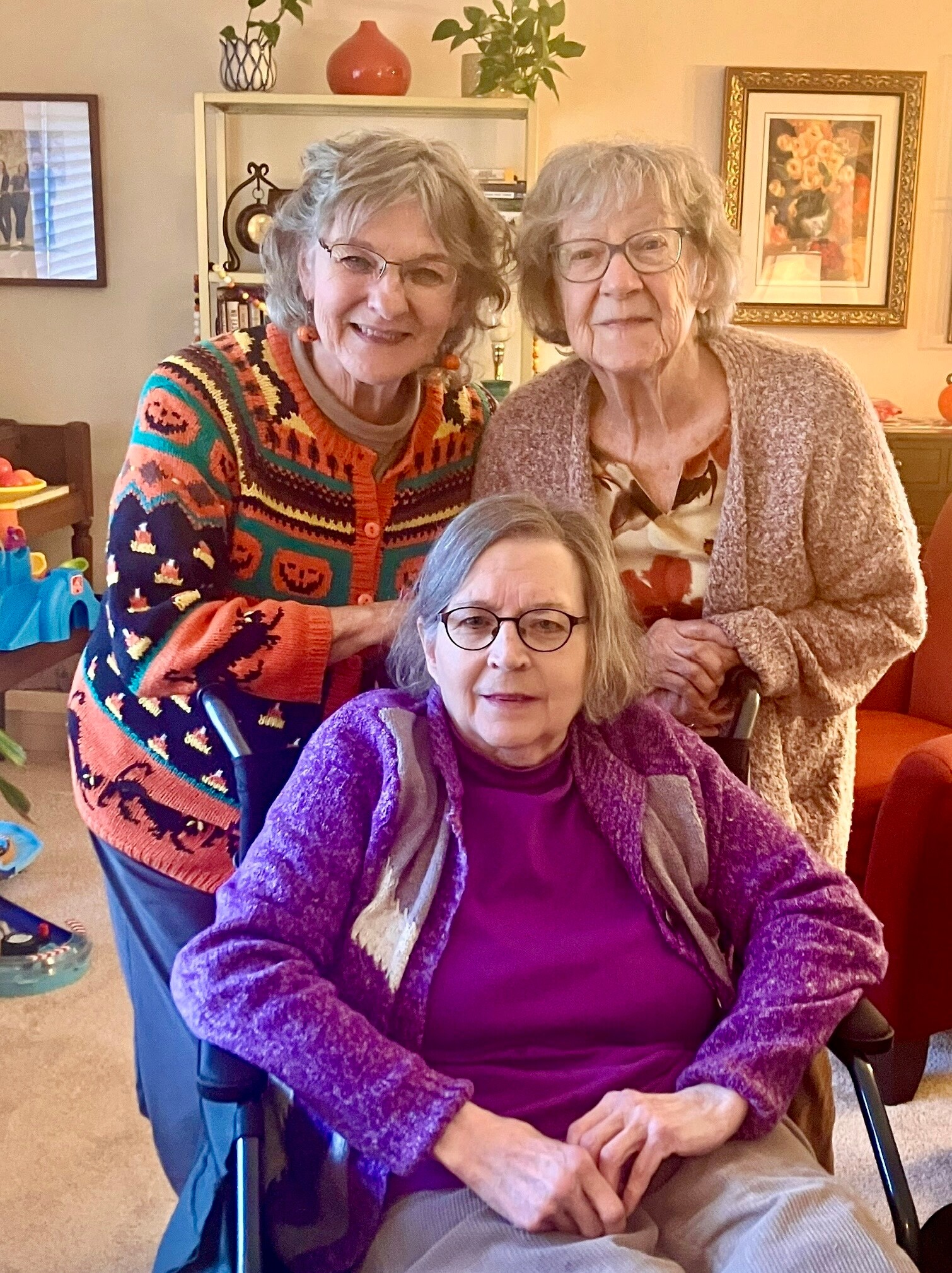 Three elderly women indoors; one seated in a wheelchair wearing purple, with two standing behind, all smiling.