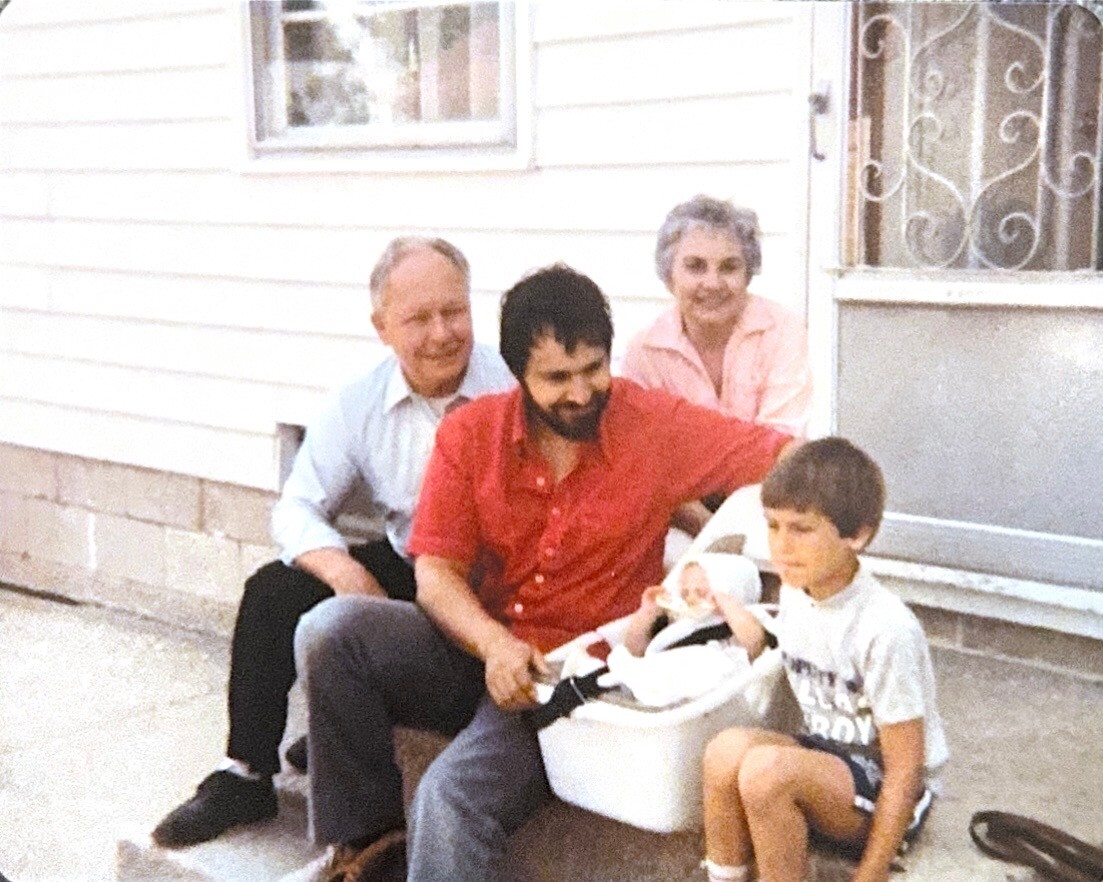 Man in a red shirt holding a baby in a car seat on a doorstep; two grandparents and a boy sit nearby.