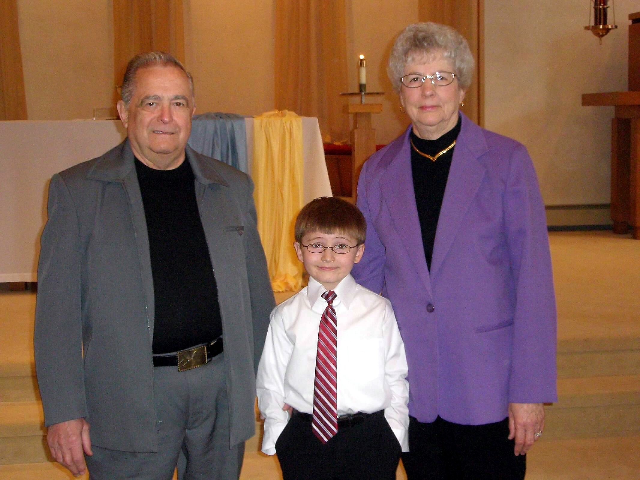 Three people posing for a photo in a church: an older man in a gray suit, a young boy in a white shirt and striped tie, and an older woman in a purple coat.
