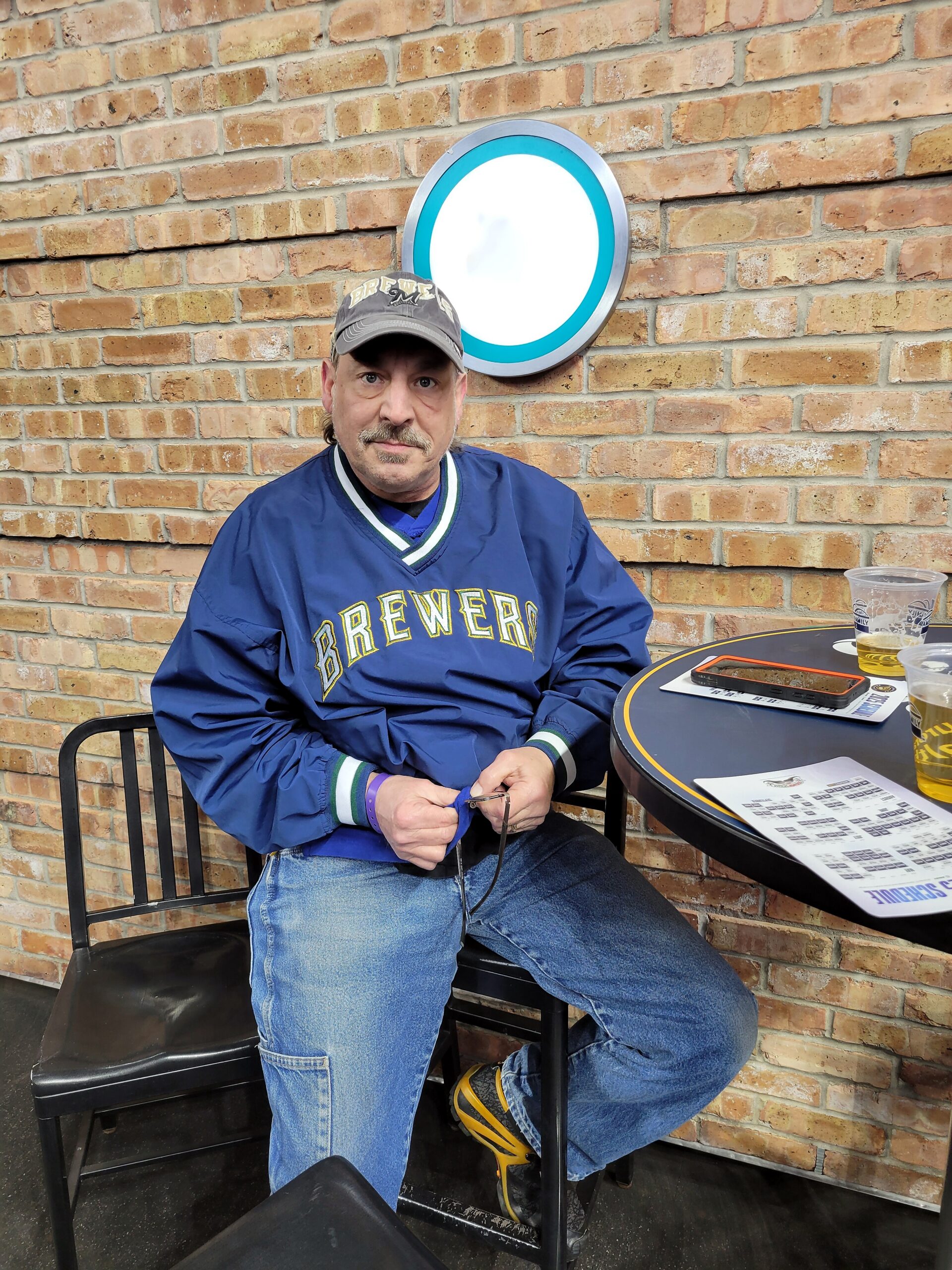 Middle-aged man in a Milwaukee Brewers jacket sits at an outdoor table against a brick wall, with drinks on the table.