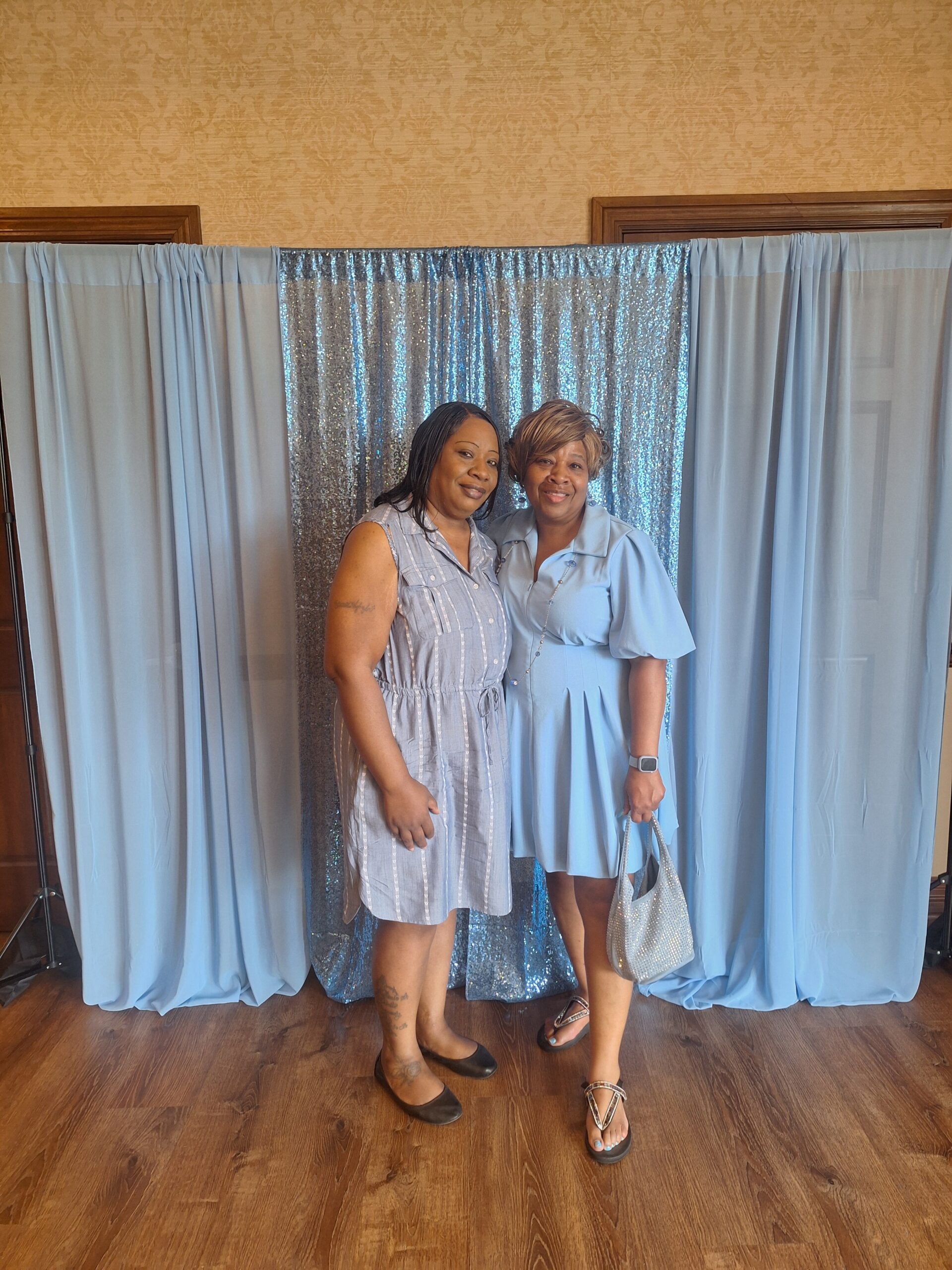 Two women smiling and posing in front of a blue sequin backdrop with light blue curtains.