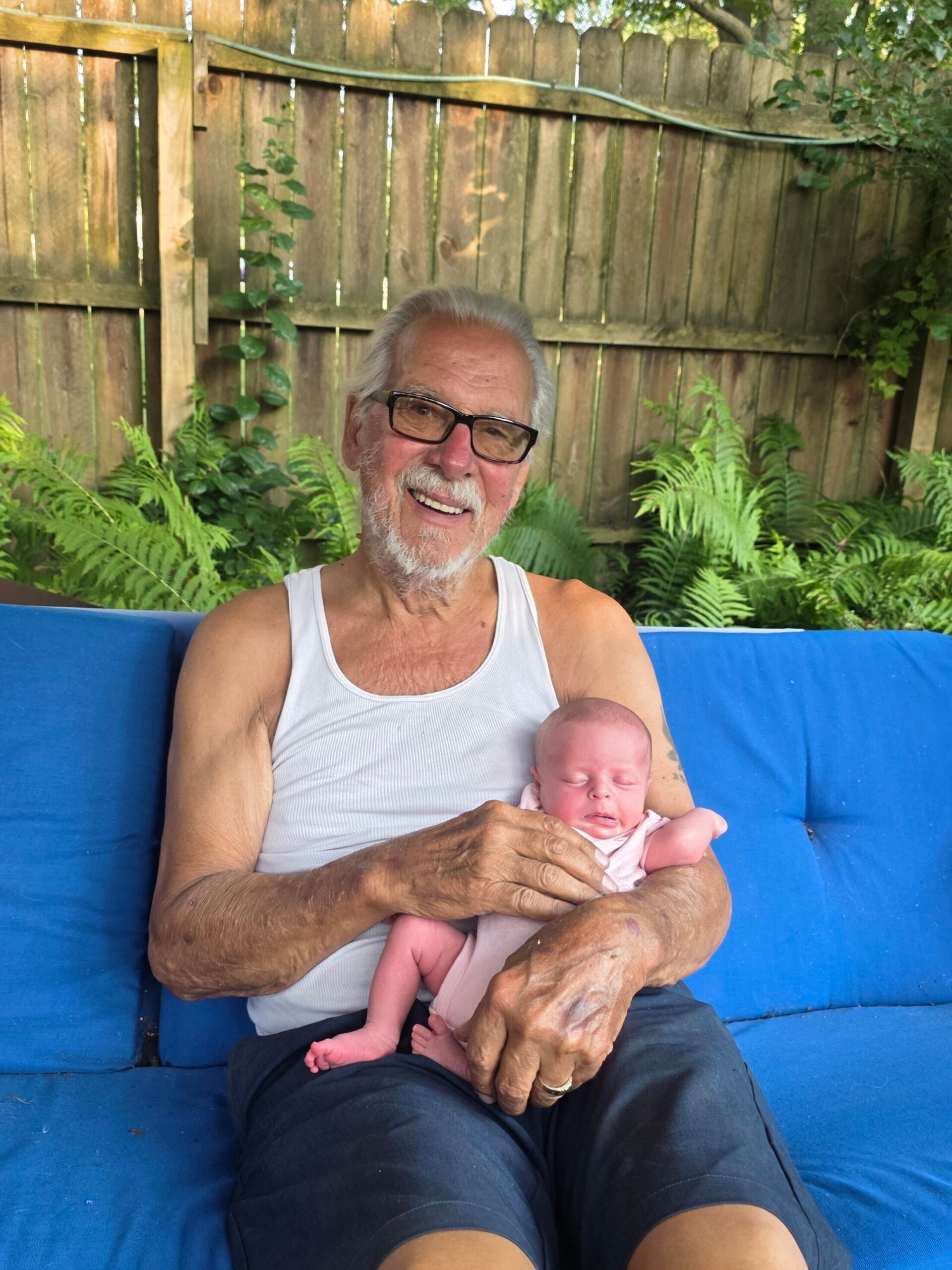 Older man with glasses smiles while sitting on a blue outdoor couch and holding a sleeping newborn baby outdoors.