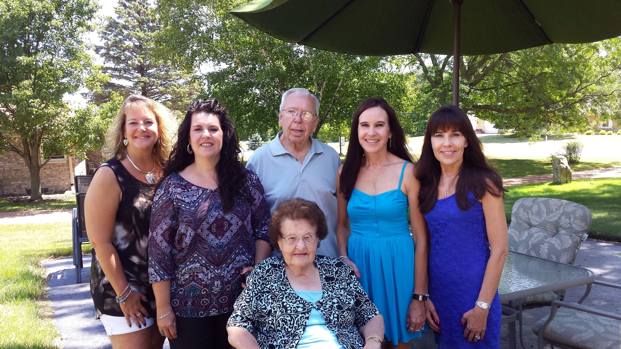 Group of six adults posing outdoors under a large umbrella, smiling in a sunny yard with trees in the background.
