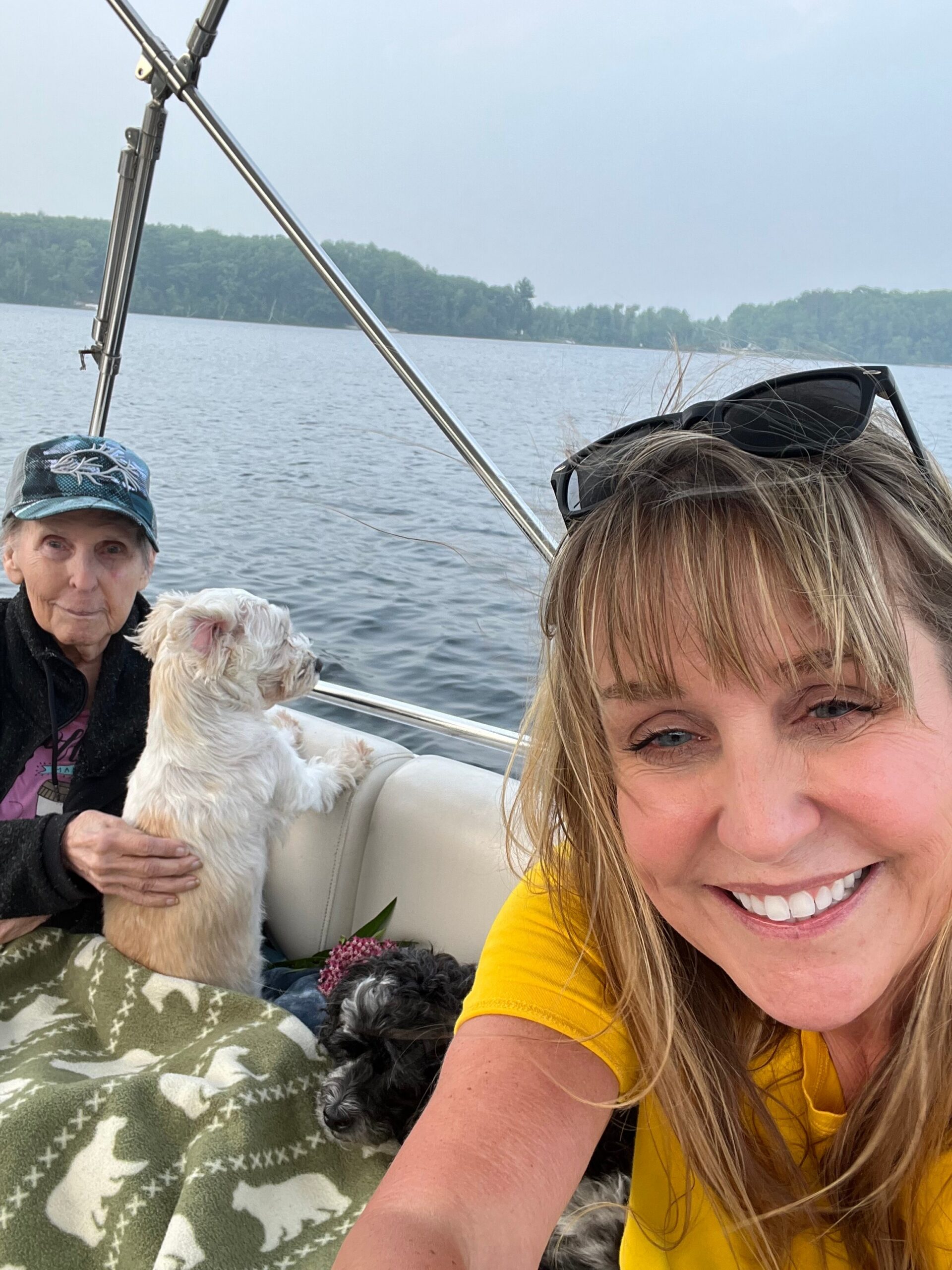 Smiling woman in a yellow shirt takes a selfie on a boat, with two dogs and a lake in the background.