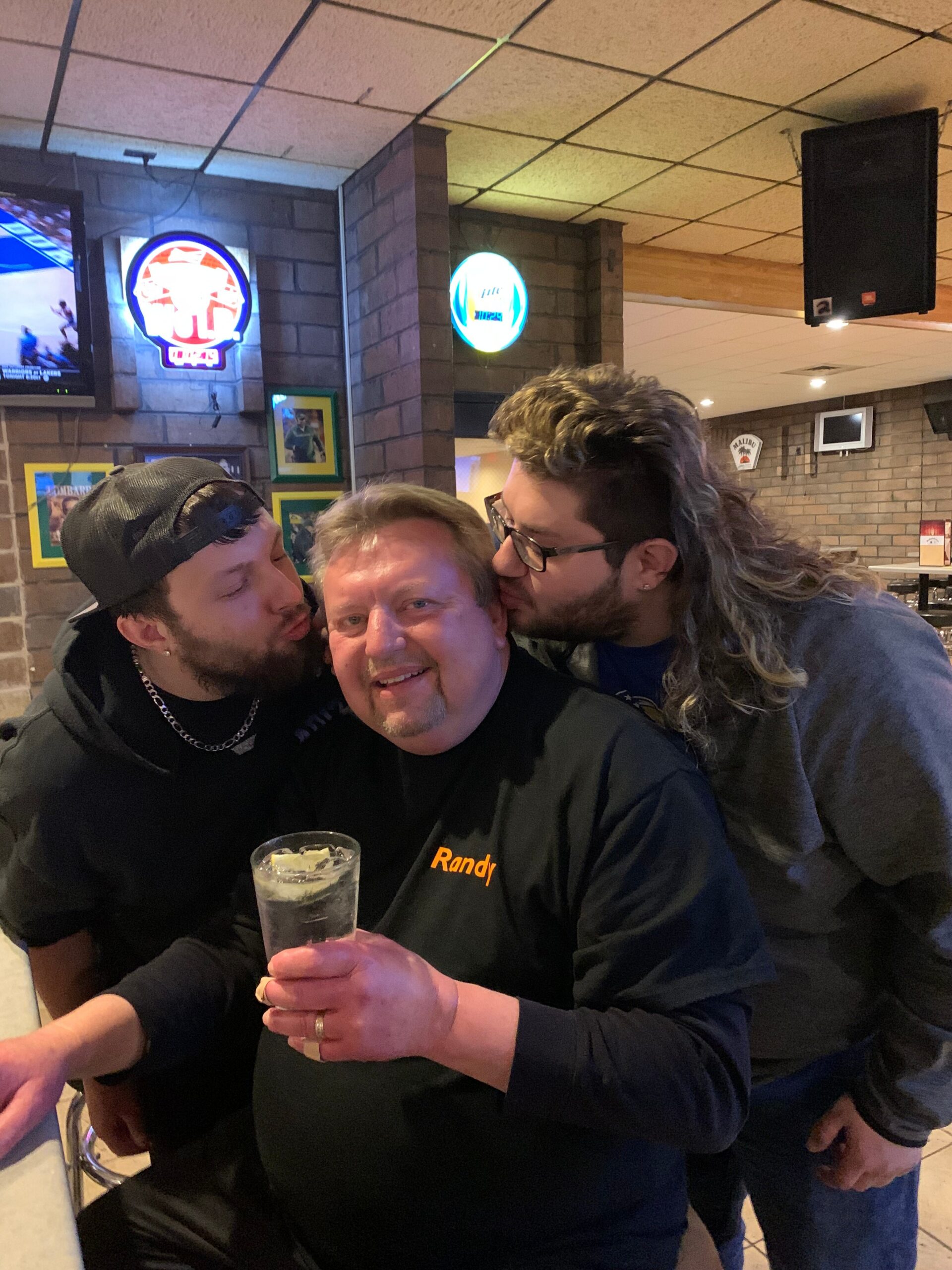 Three men in a bar pose for a photo; center man wears a black shirt with 'Randy' and holds a drink while two friends kiss his cheeks from either side.