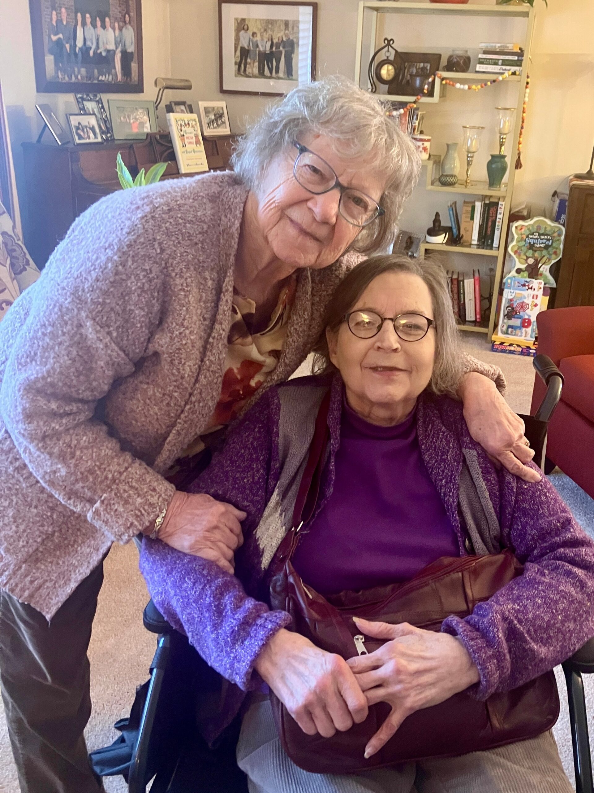 Two elderly women in a cozy living room; one stands and hugs the other in a wheelchair, both smiling.
