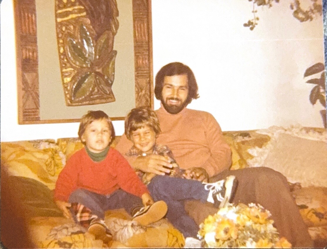 Father with two young children sitting on a floral sofa in a living room, the man in a rust-colored shirt holding a bottle and smiling.