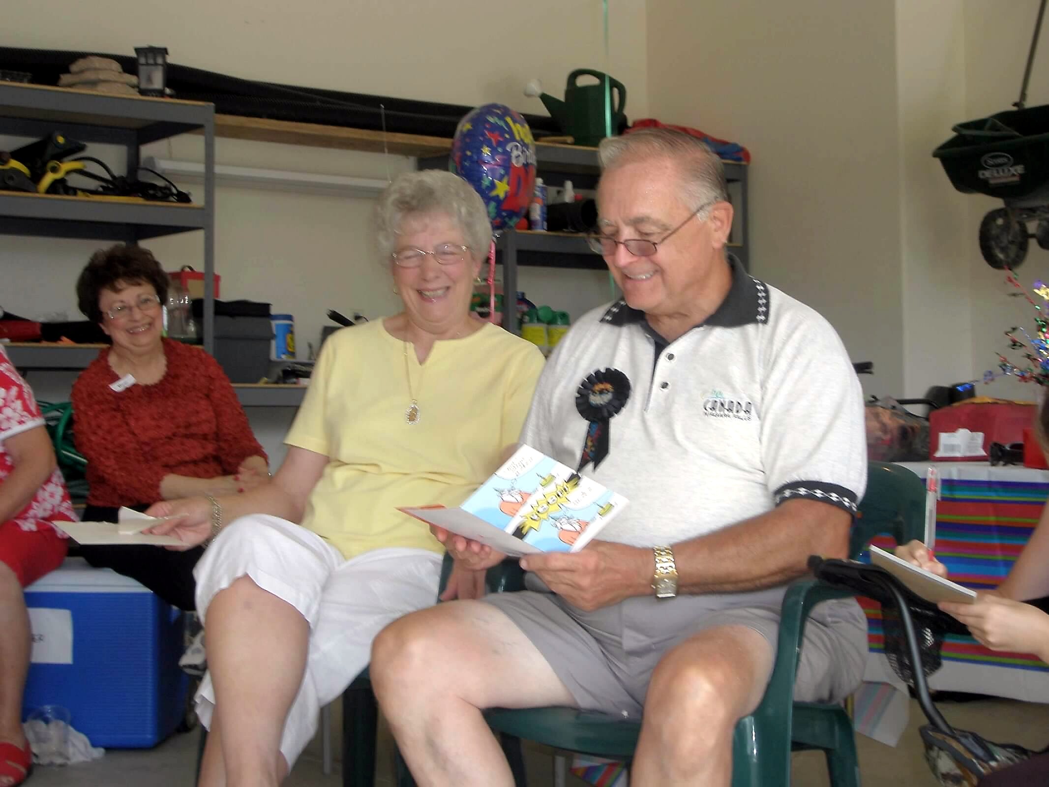 Smiling elderly couple reading a card at a celebration, with balloons in the background in a garage or community room.