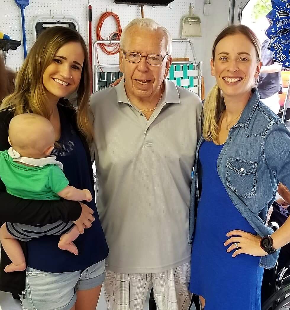 Three adults and a baby posing in a workshop; woman on the left holds a baby, an elderly man stands center, and a woman on the right wears a denim jacket.