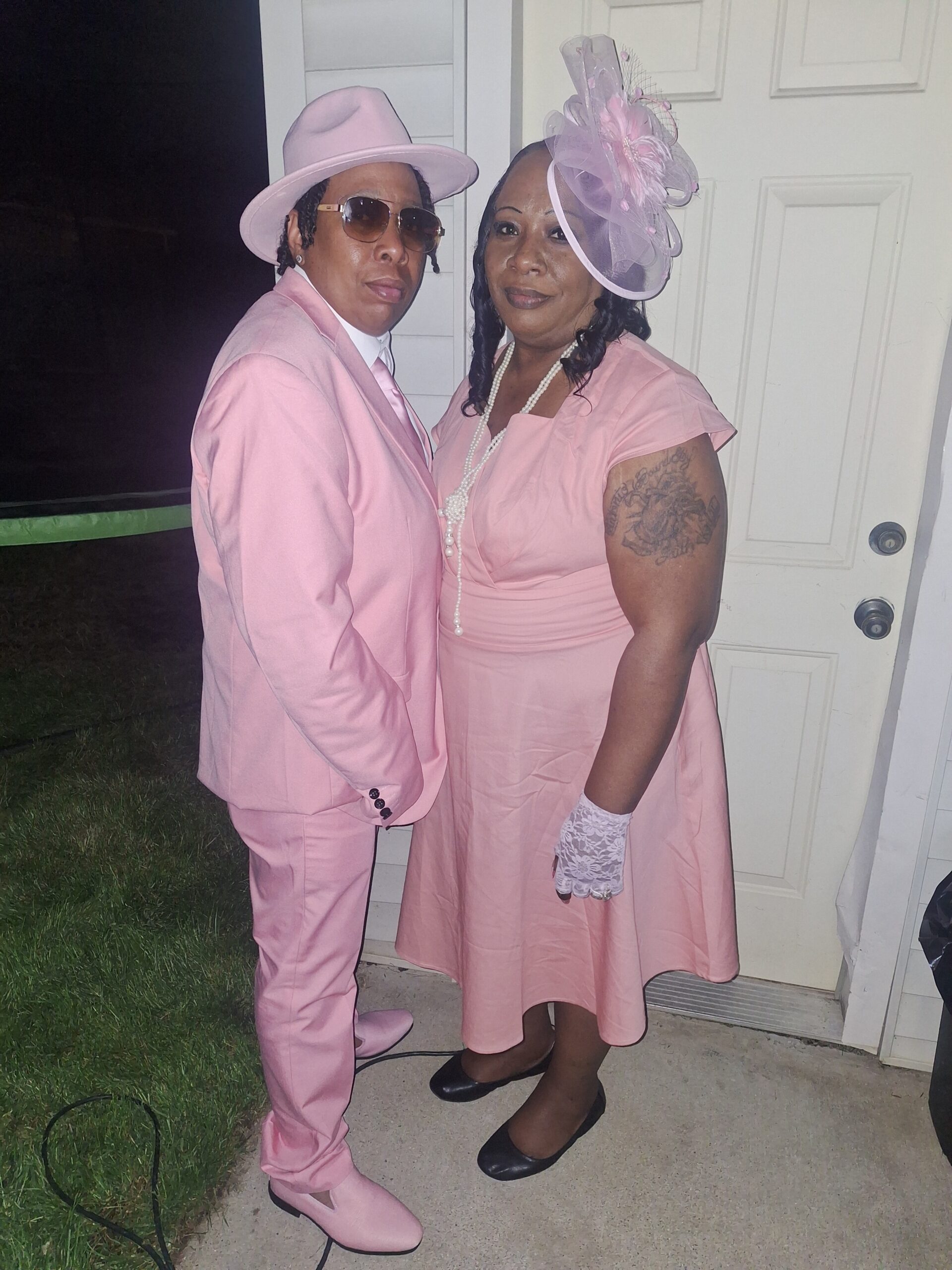 Two people posing together in matching pink outfits: a man in a pink suit with a pink hat and sunglasses and a woman in a pink dress with a decorative pink hat, pearl necklace, and white lace gloves standing by a door outdoors.