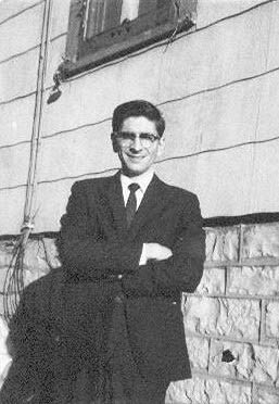 Young man in a suit and tie stands with arms crossed beside a stone wall, with a window above him.
