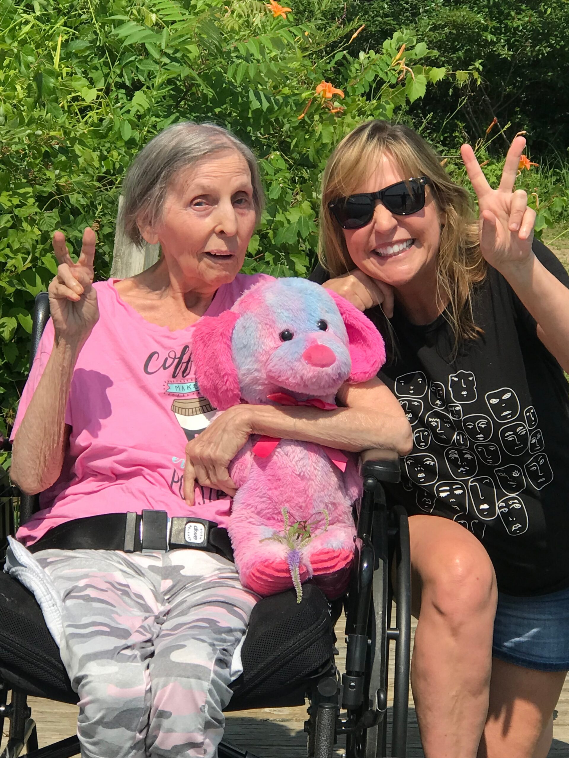 Two women outdoors posing with peace signs; one elderly woman in a pink shirt in a wheelchair holding a pink plush toy, with a younger woman in sunglasses beside her behind green foliage.