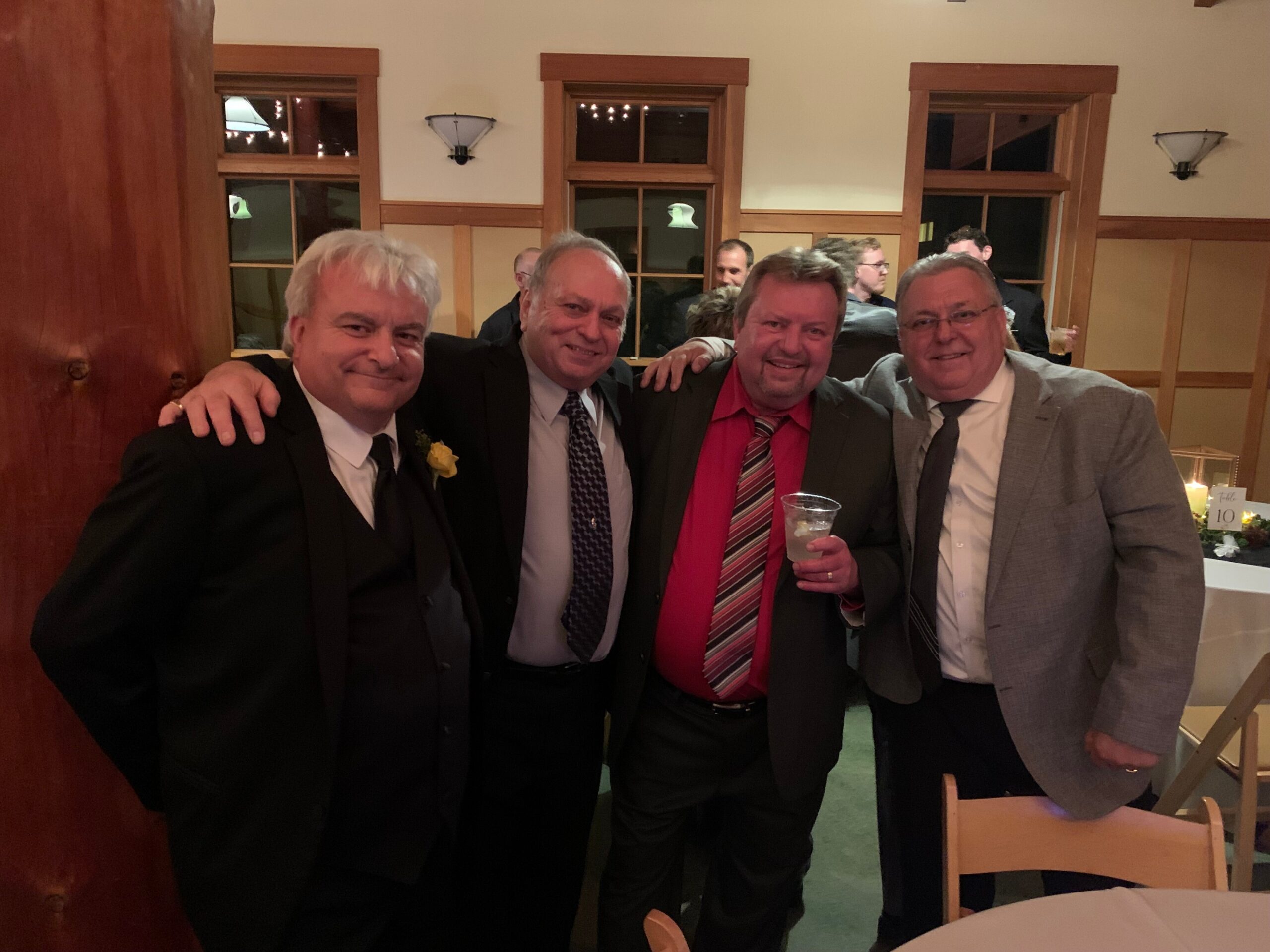 Four men in suits posing together at a social event, smiling for the camera in a warmly lit room.