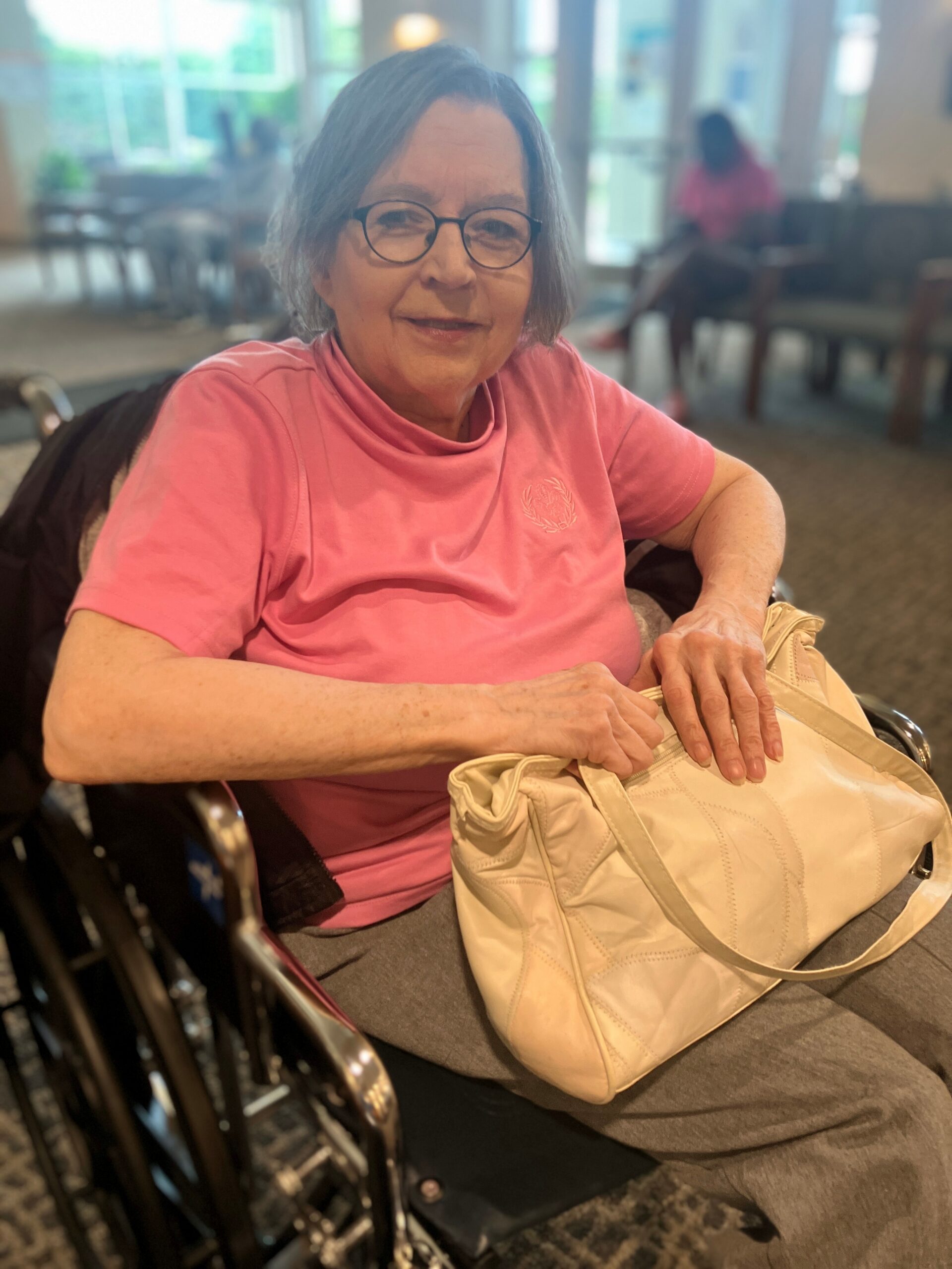 Older woman with short gray hair and glasses sits in a wheelchair, wearing a pink shirt and holding a beige handbag on her lap.