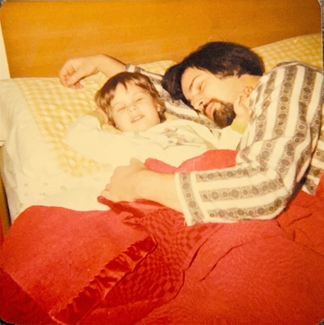 Father and child asleep together in bed, smiling as they cuddle under a red blanket and patterned coverlet ideal for a cozy, family moment (vintage photo).] ,