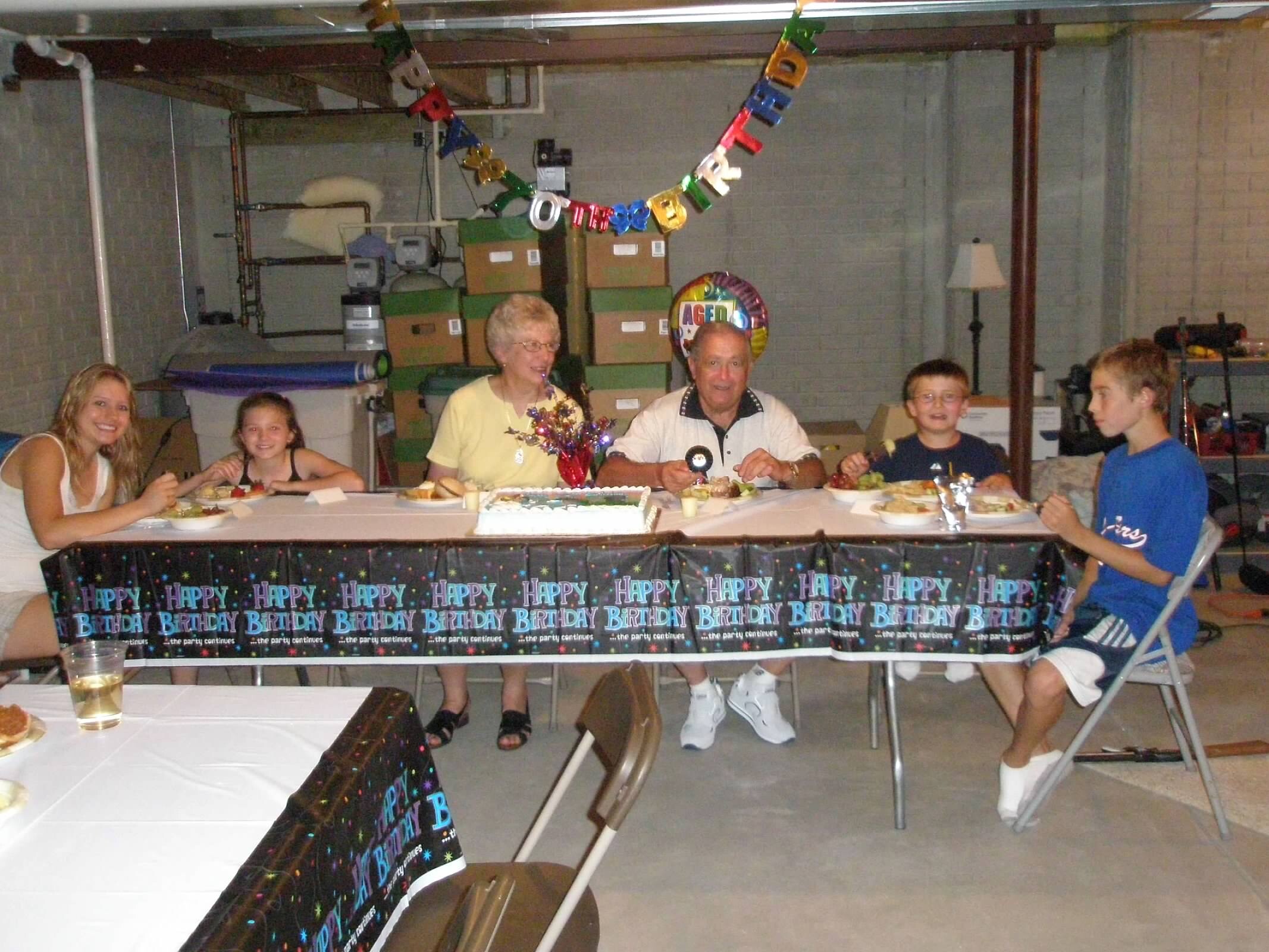 Family birthday gathering at a long table in a basement, with a colorful 'Happy Birthday' banner along the edge and a cake in the center as relatives enjoy a meal.