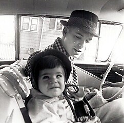 Man wearing a hat sits in the driver's seat of a vintage car with a smiling toddler in the front passenger seat in a black-and-white photo.