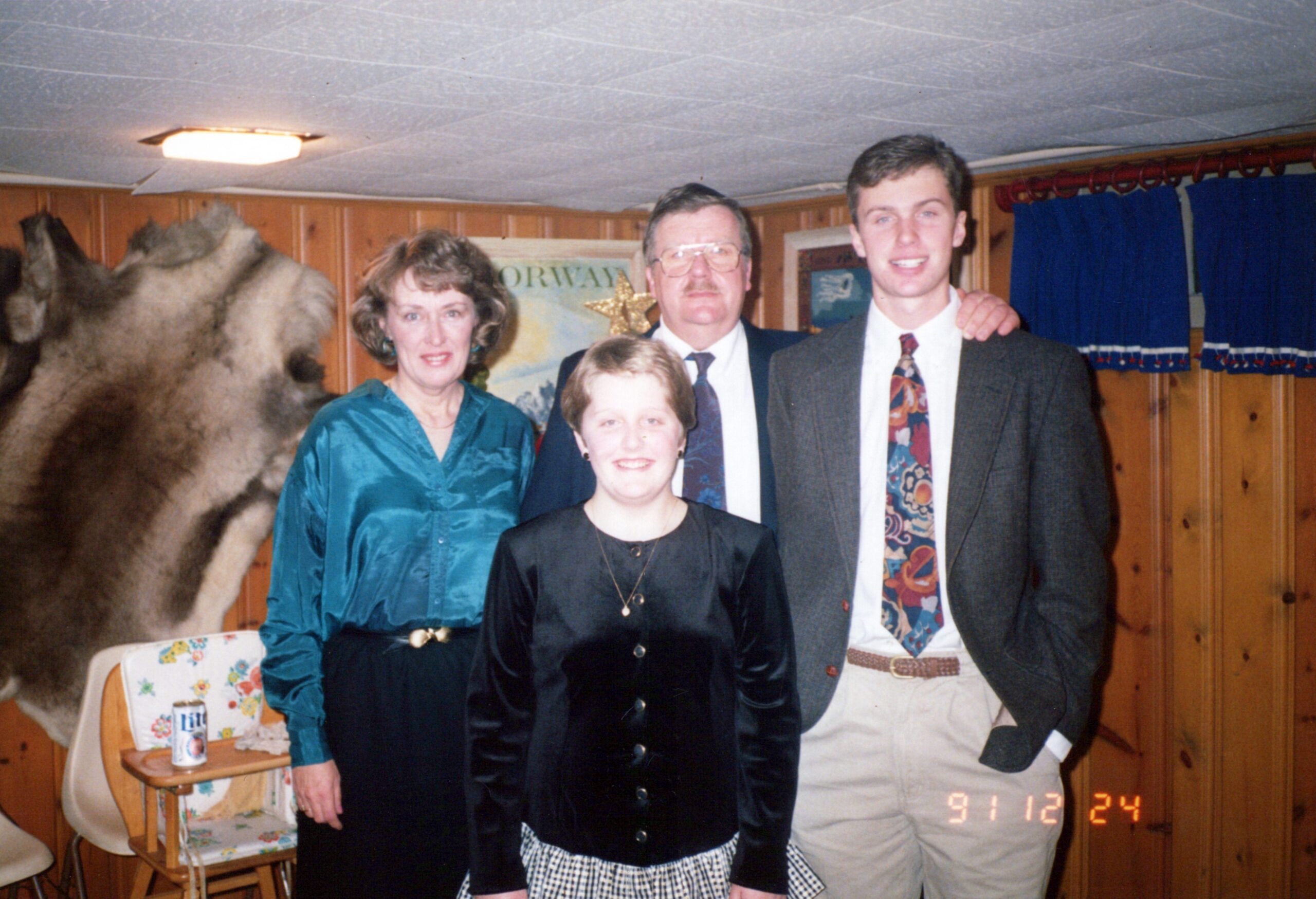 Four family members pose together in a wood-paneled room with a mounted animal hide in the background; a woman in teal, a girl in black, and two men in formal attire smile for the photo.