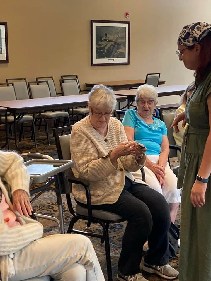 An elderly woman in a beige cardigan sits in a chair, looking at a small device in her hands, as another senior woman and a caregiver stand nearby in a communal room with tables and chairs.