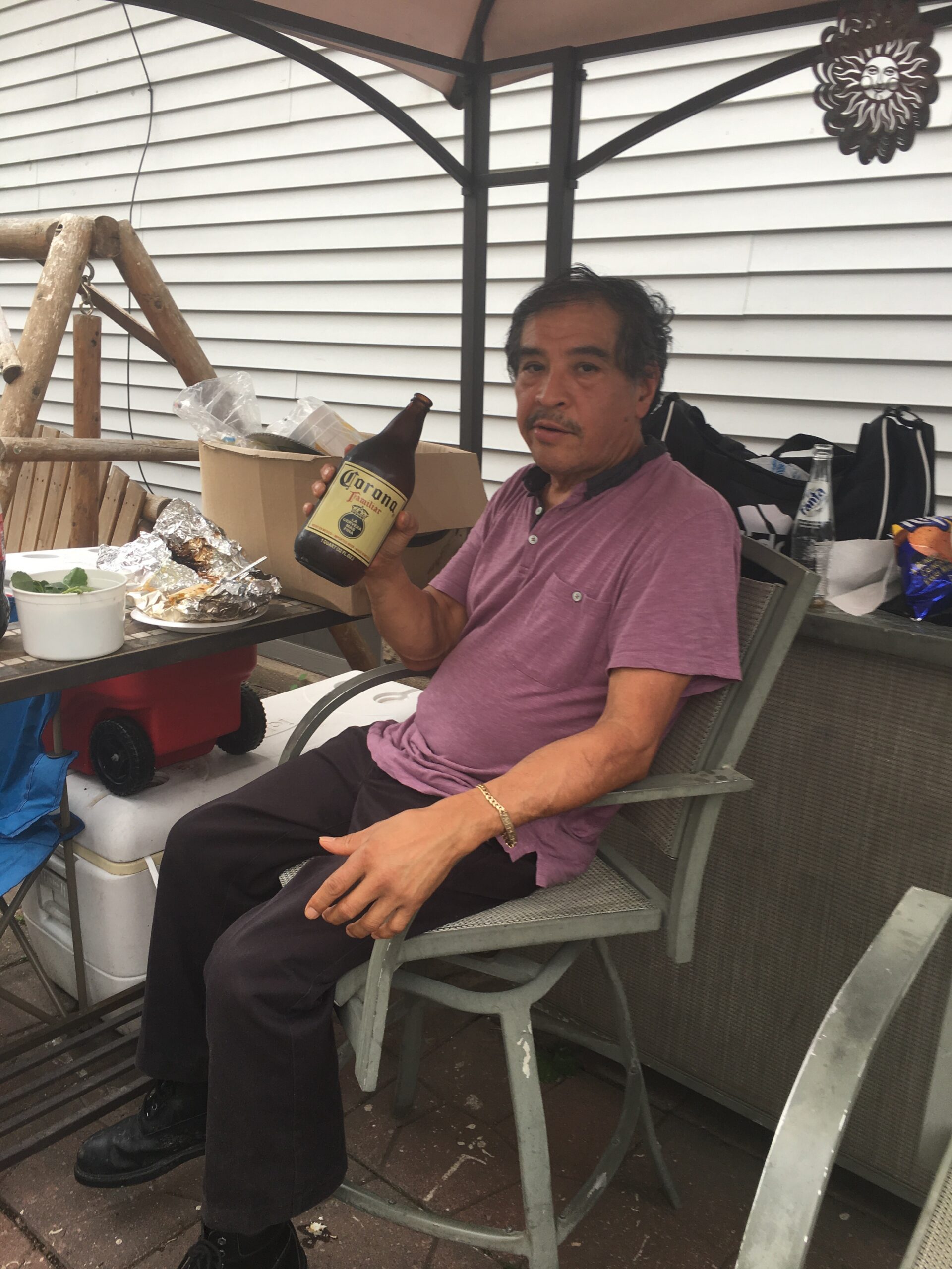 Man in a pink polo shirt sitting on a patio chair, holding a Corona beer bottle with foil-wrapped food on the table nearby.