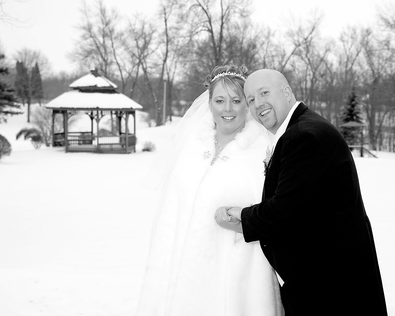 Bride and groom pose together outdoors in a snowy yard, the bride in a white fur-trimmed gown and veil, smiling beside the man in a dark coat. Behind them is a wooden gazebo dusted with snow.] ,