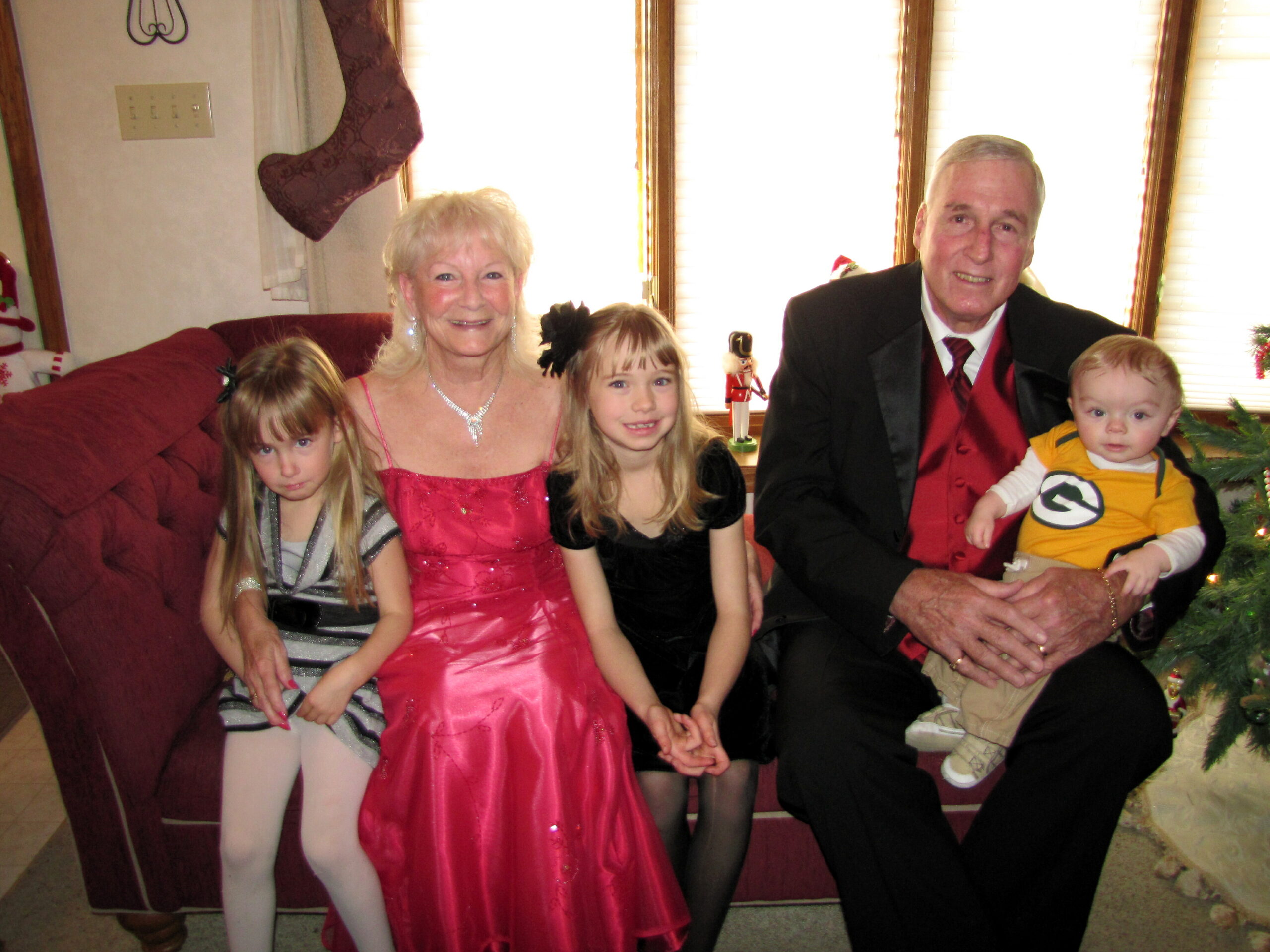 Family group photo on a red sofa in a Christmas living room, smiling adults and children dressed up for the holidays with a baby on the man's lap.