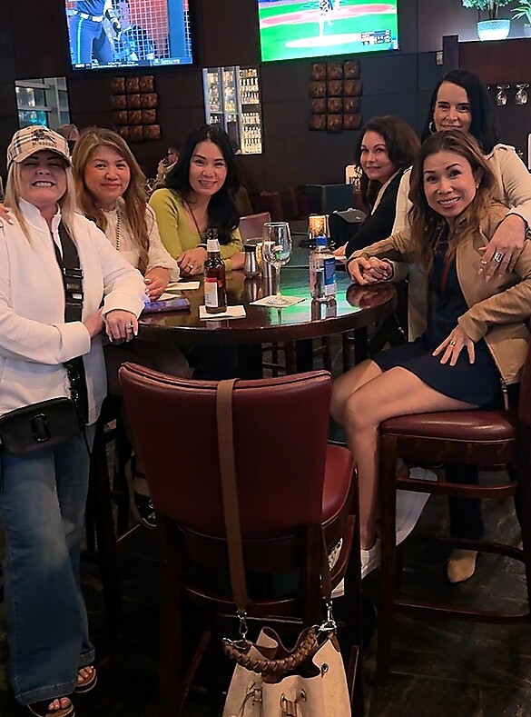 Six women sit around a round bar table in a lounge, smiling with drinks and a bottle on the table, TV screens in the background.