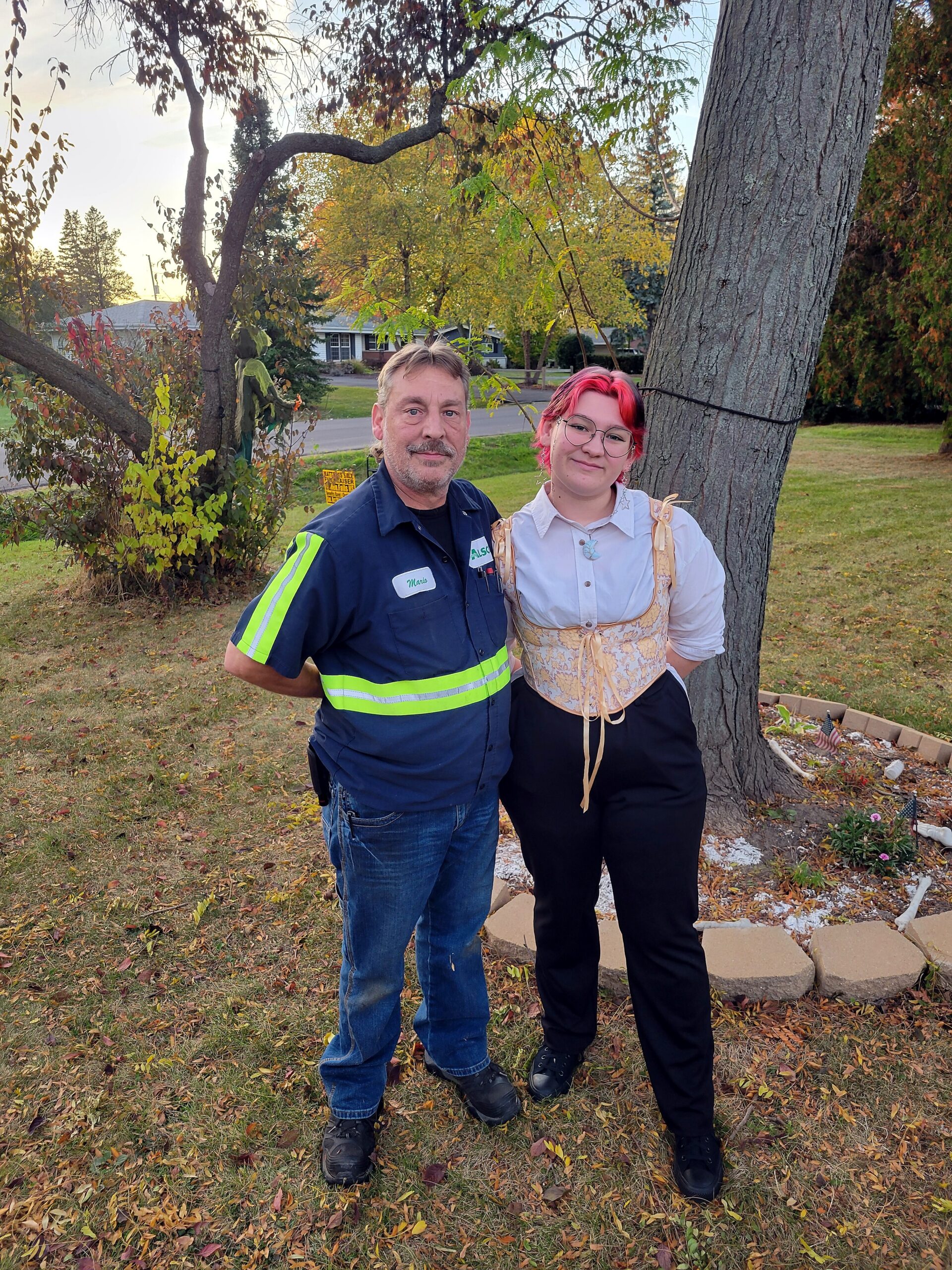 Man in a blue work shirt with neon stripes and a woman with pink hair pose together in a fall yard by a tree, smiling at the camera.