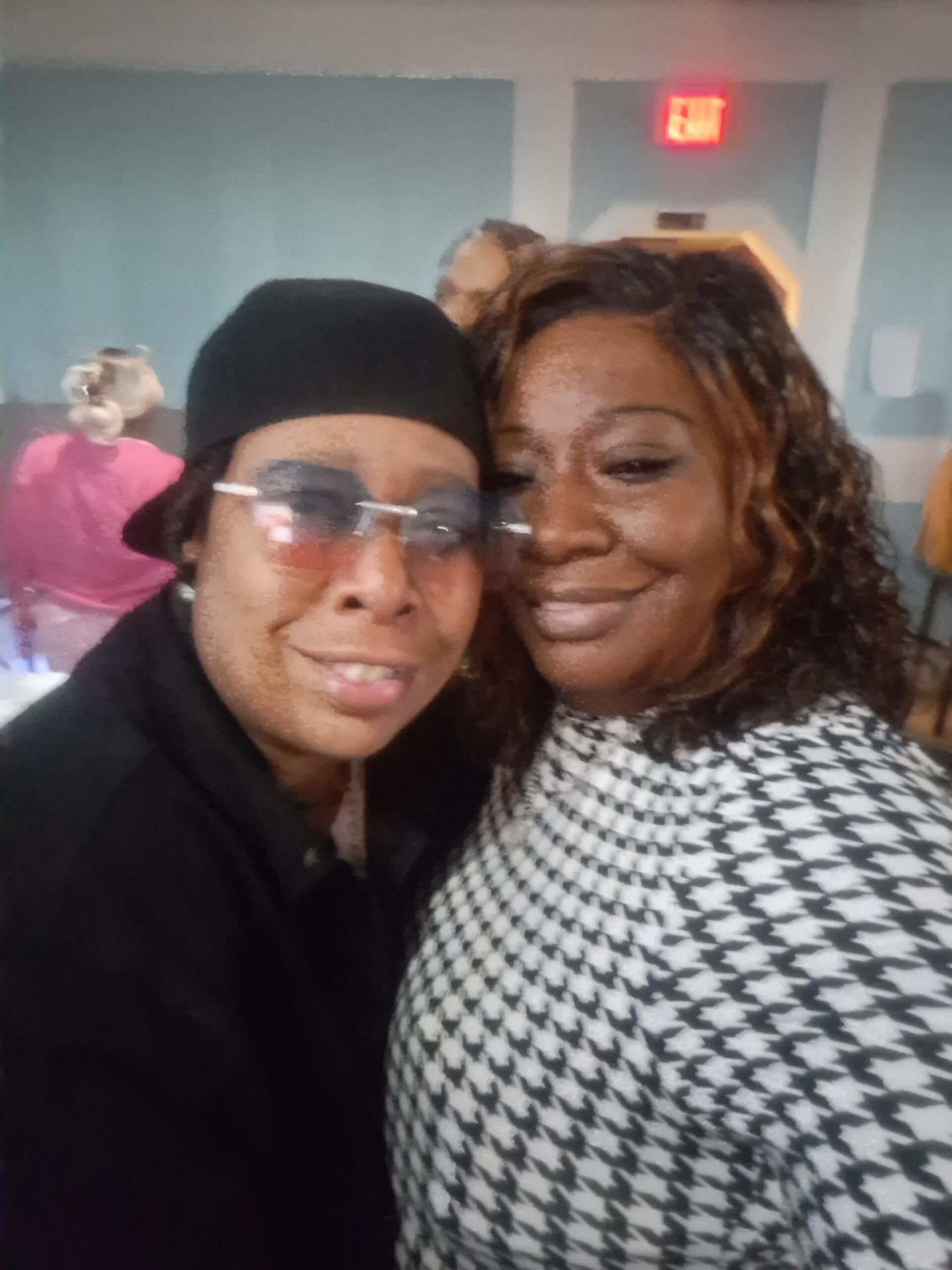 Two friends pose for a close selfie indoors, smiling at the camera with one wearing a black beanie and glasses and the other in a houndstooth top, background shows blue walls and an illuminated exit sign.