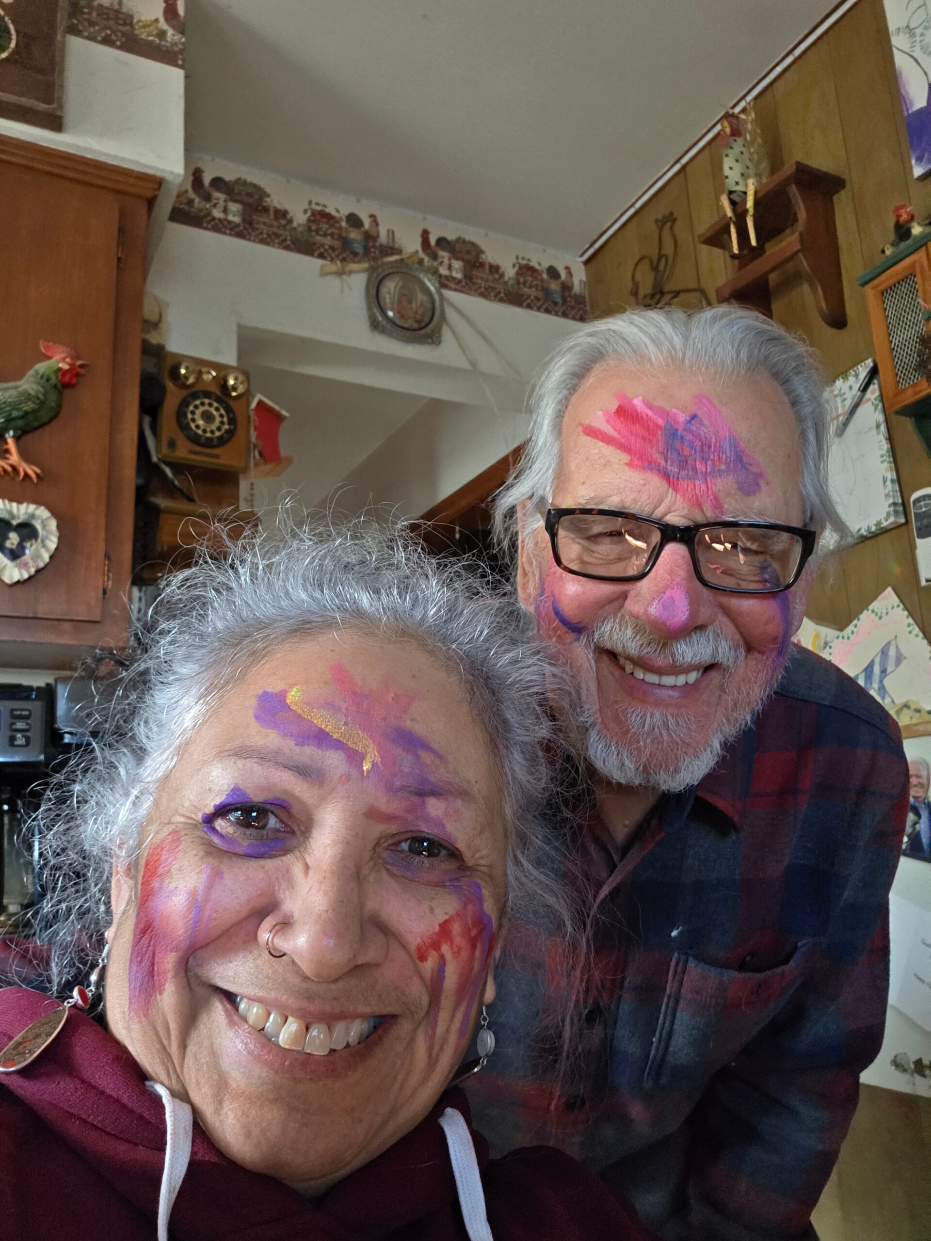 Smiling elderly couple with bright face paint posing in a home kitchen. The woman in front has purple and pink paint and the man behind has matching colors.