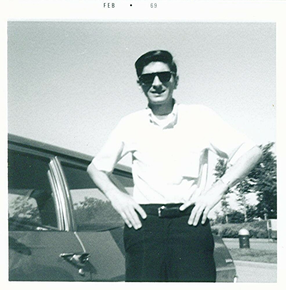 Man in a white shirt and dark pants wearing sunglasses, standing with hands on hips beside a car (Feb 1969).