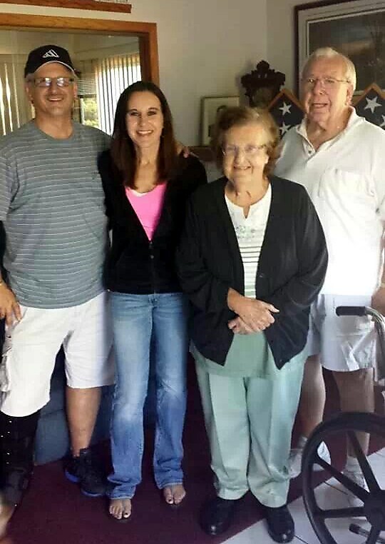 Four people pose for a photo in a living room: a man in a striped shirt, a young woman in pink, an elderly woman in a cardigan, and an older man in a white shirt, with a wheelchair nearby.