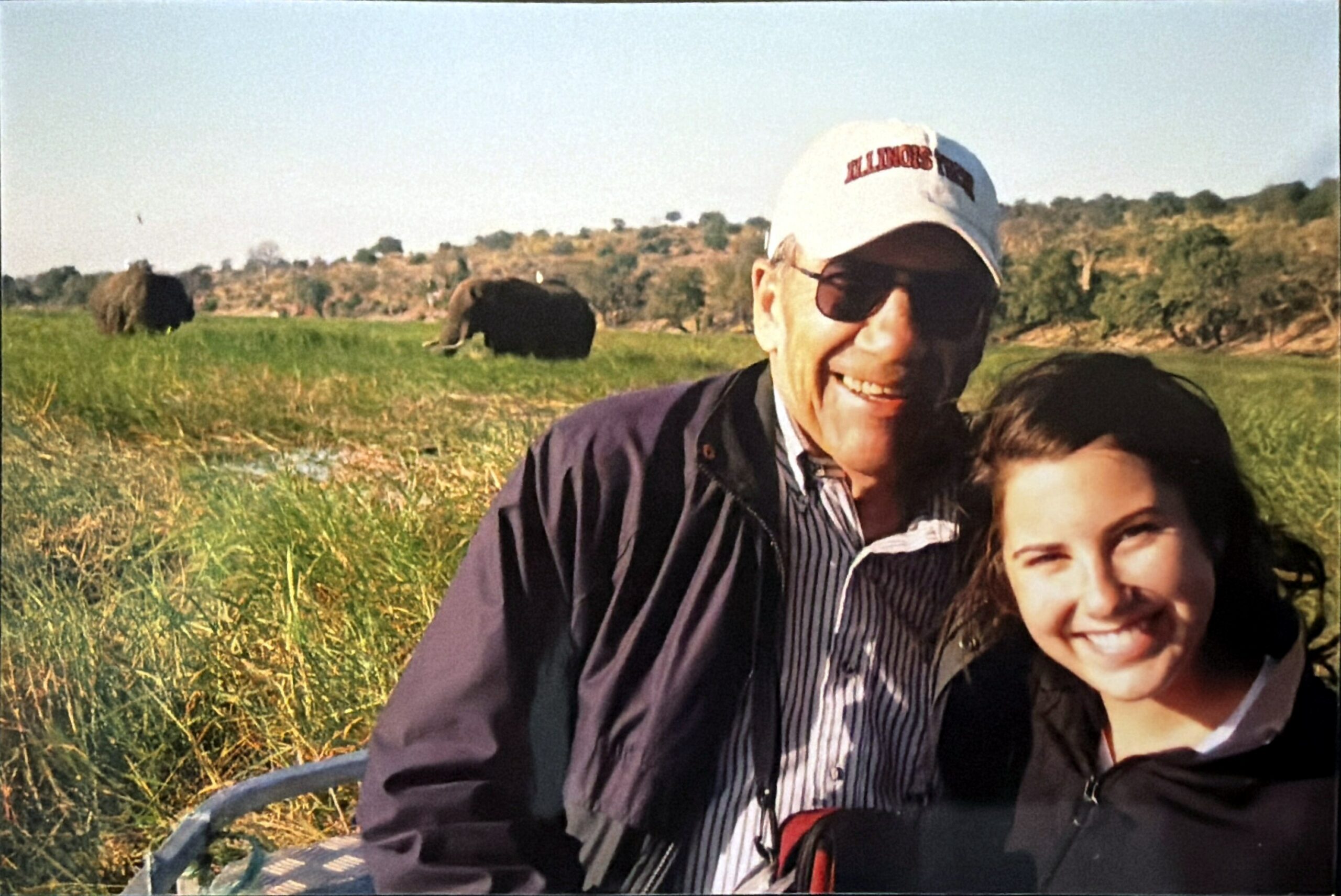 Two people smiling for a selfie in a grassy field with elephants grazing in the distance in the background.