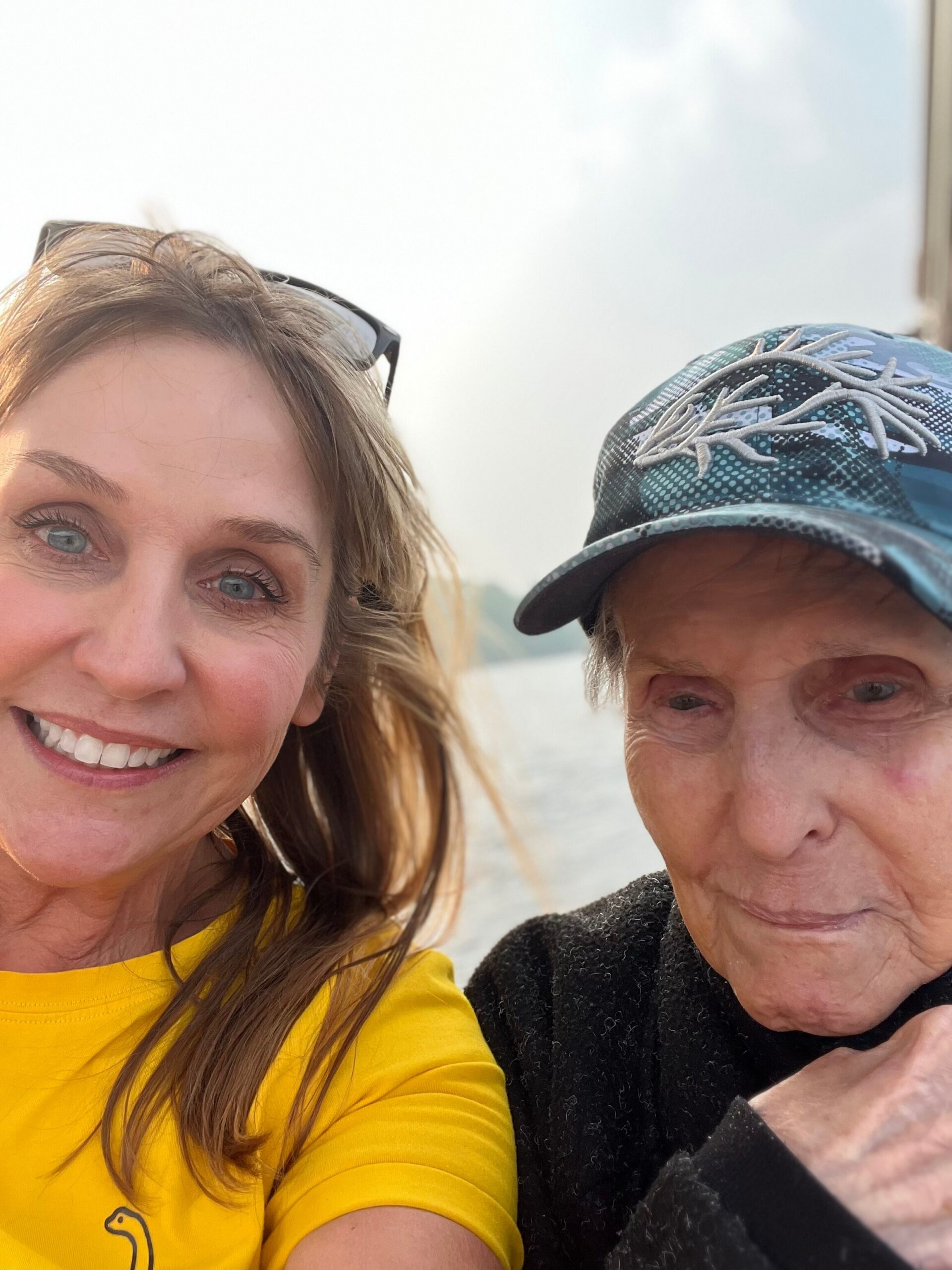 Two people posing for a selfie outdoors, smiling at the camera with water in the background.