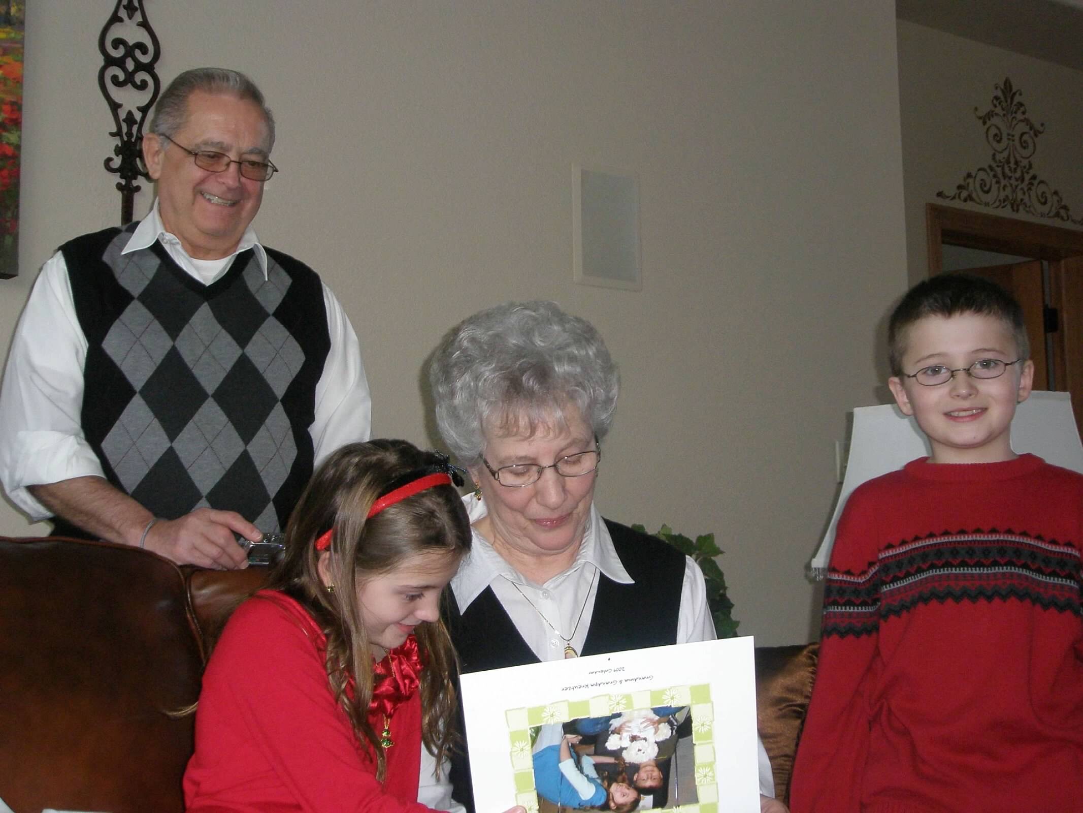 Older man in a gray argyle sweater vest and glasses smiles as children hold up a large photo collage indoors with decorative wall art nearby.