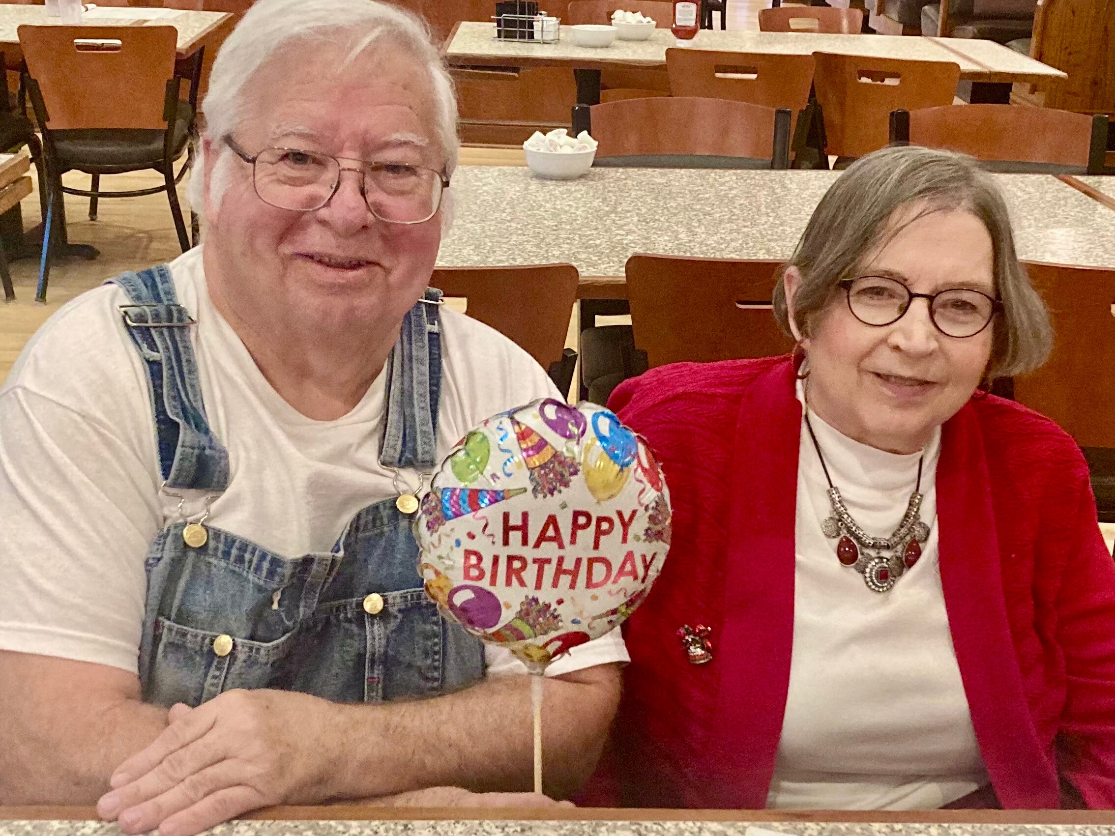 Two elderly friends at a restaurant table, a colorful birthday balloon between them reads 'HAPPY BIRTHDAY'.