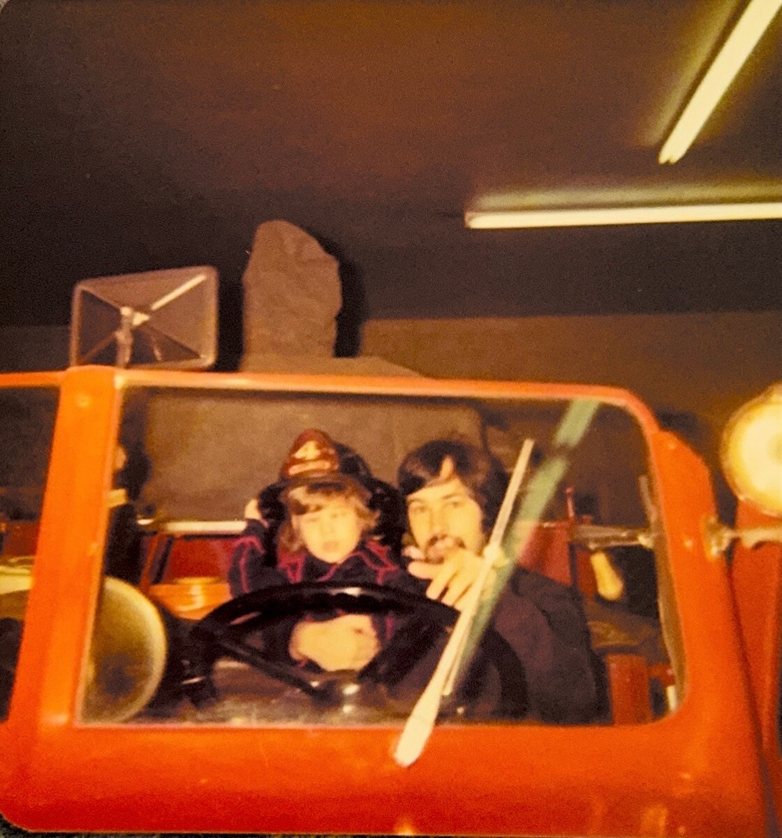 Two people sit in a small red vintage car, posing for a photo indoors behind the windshield.