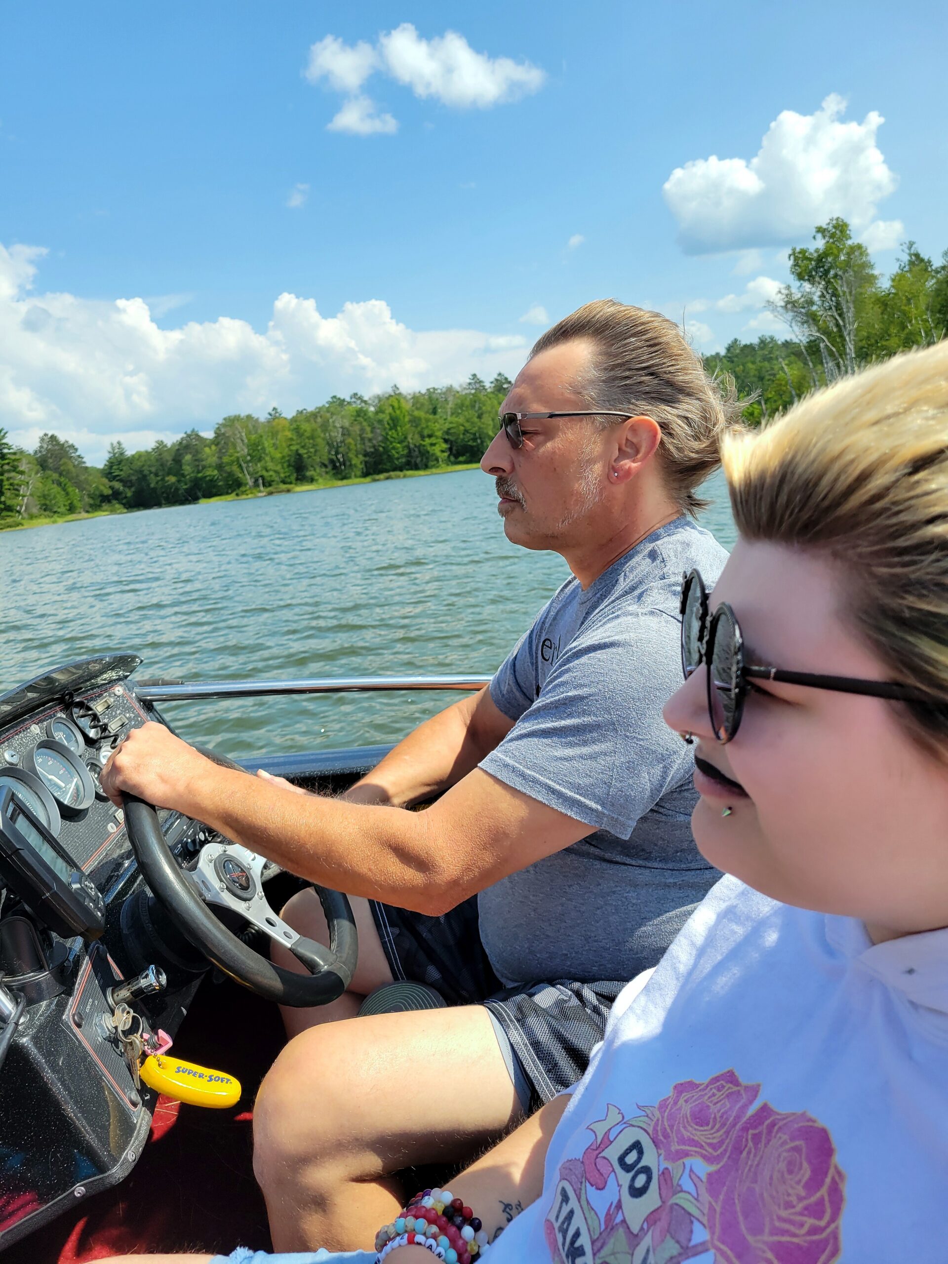 Man wearing sunglasses steers a motorboat on a lake with a forested shoreline in the background; a woman in the passenger seat wears sunglasses in the foreground.
