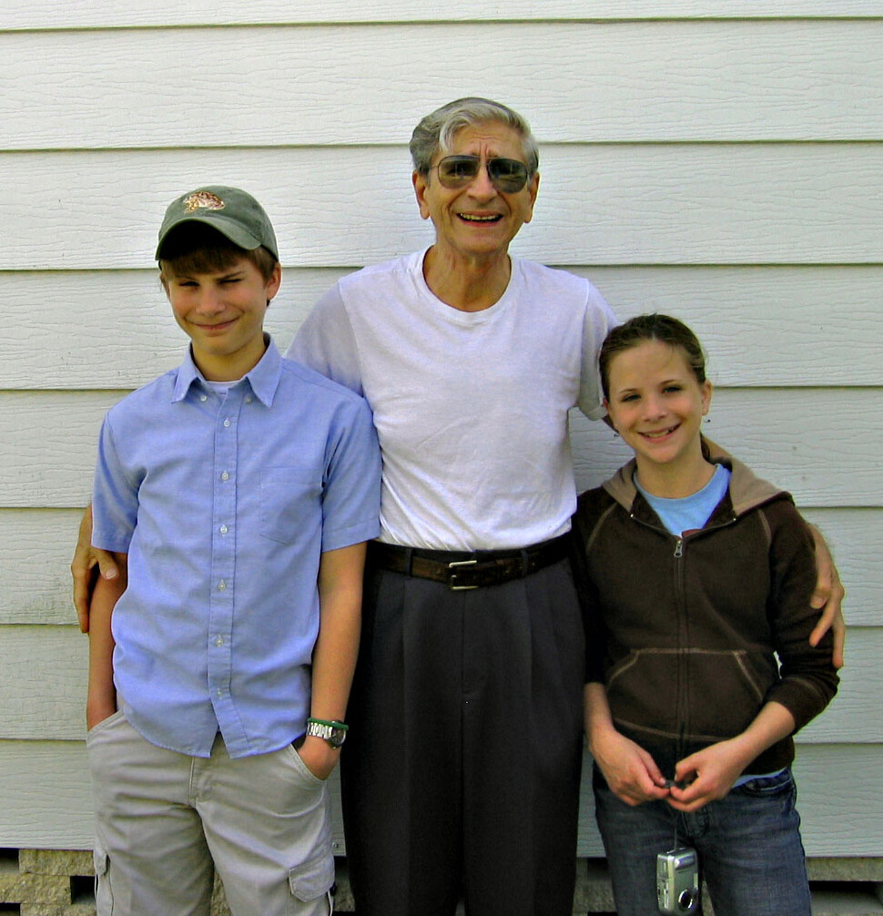 Older man wearing a white T-shirt and sunglasses stands with a boy in a blue shirt and a girl in a brown hoodie outside a light-gray siding wall.