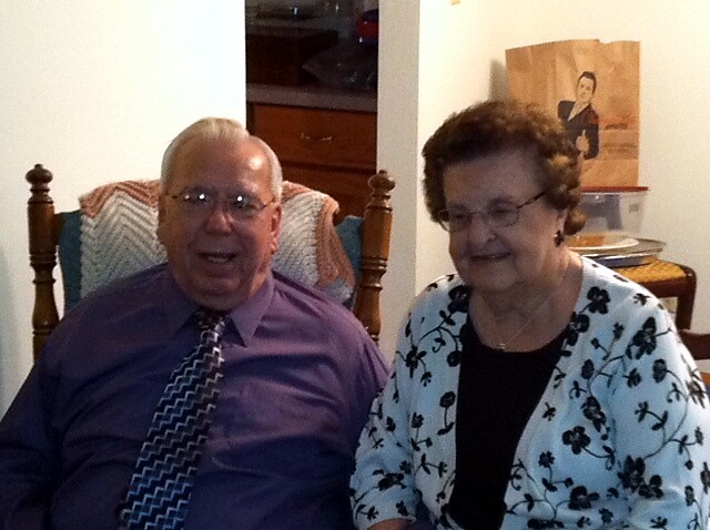 Elderly man in a purple shirt with a patterned tie sits next to an elderly woman in a black top and white floral cardigan, smiling in a living room with wooden chairs.