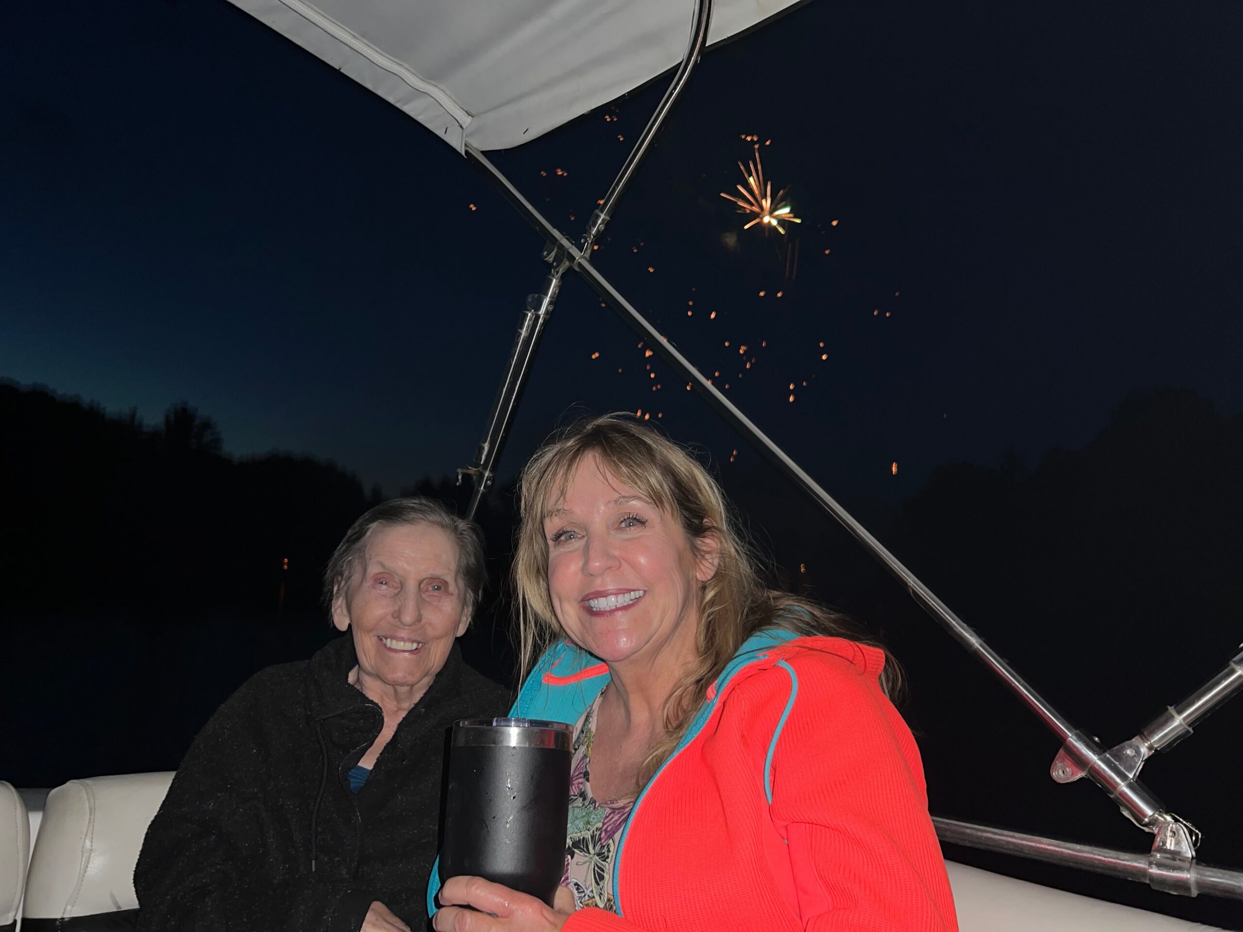 Two women smiling on a boat at night, holding drinks, with fireworks in the dark sky behind them.