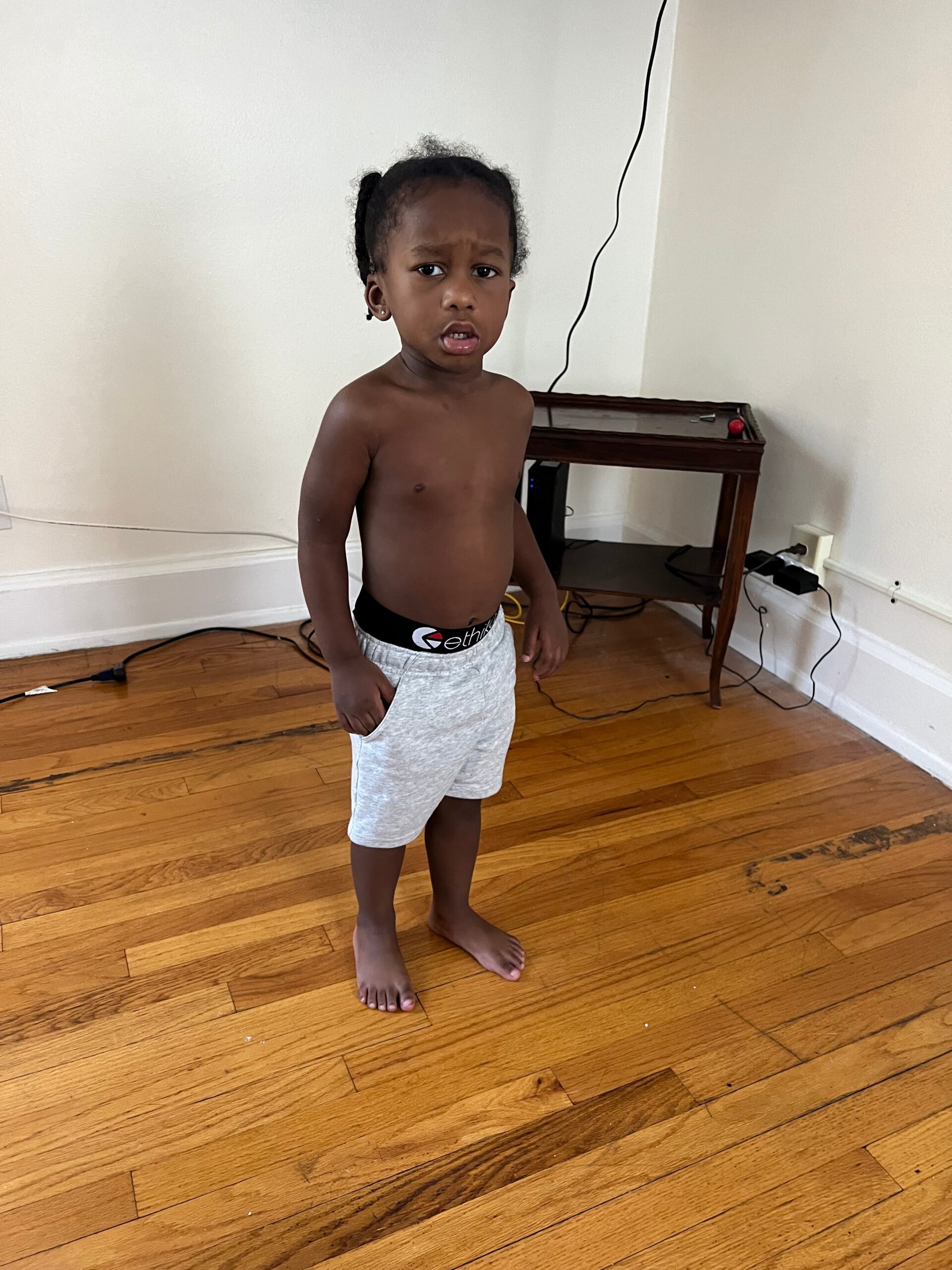 Young child standing barefoot indoors, shirtless, wearing gray shorts, looking at the camera with a puzzled expression.