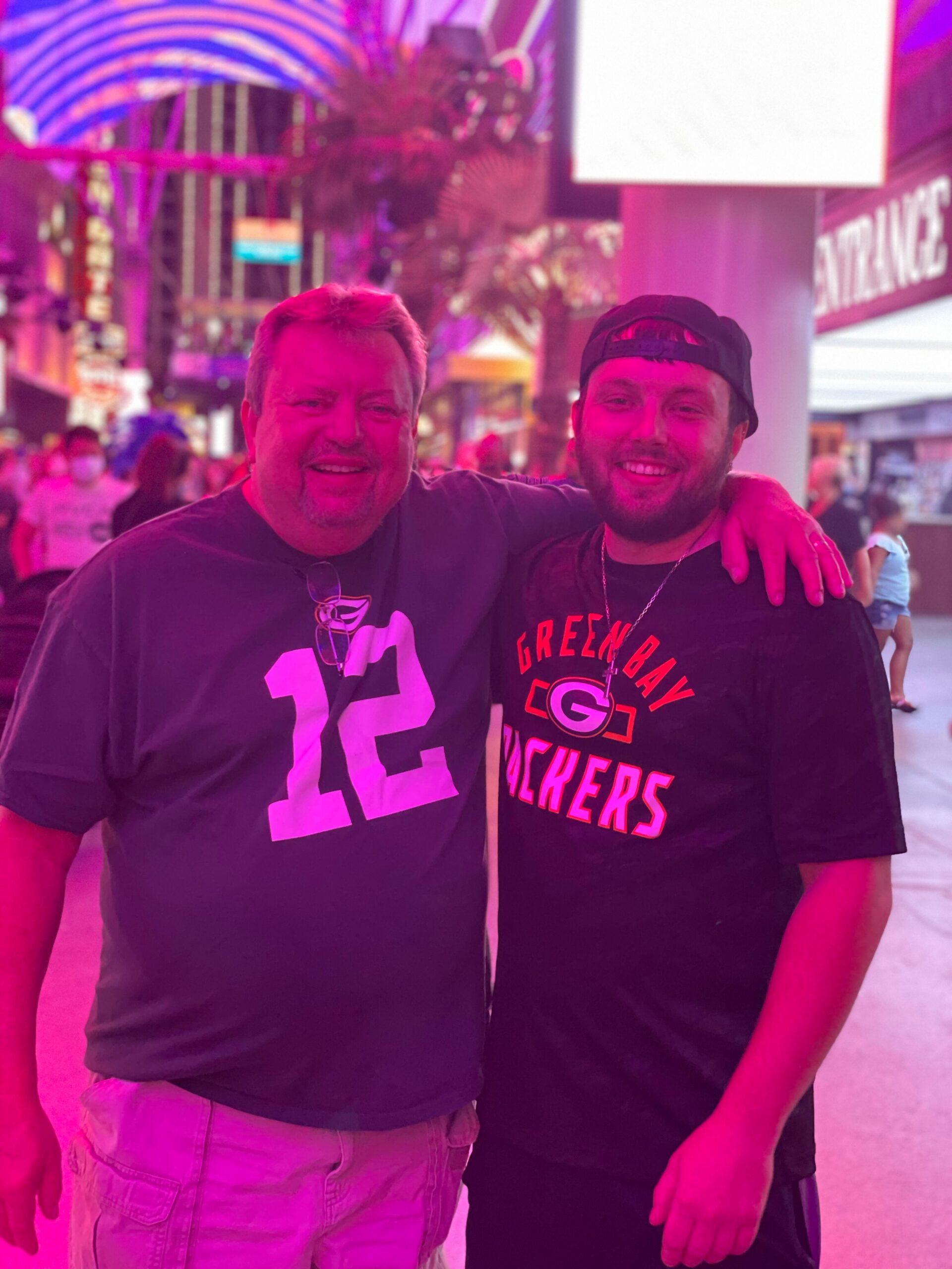 Two men smiling and posing with arms around each other at a neon-lit street event.
