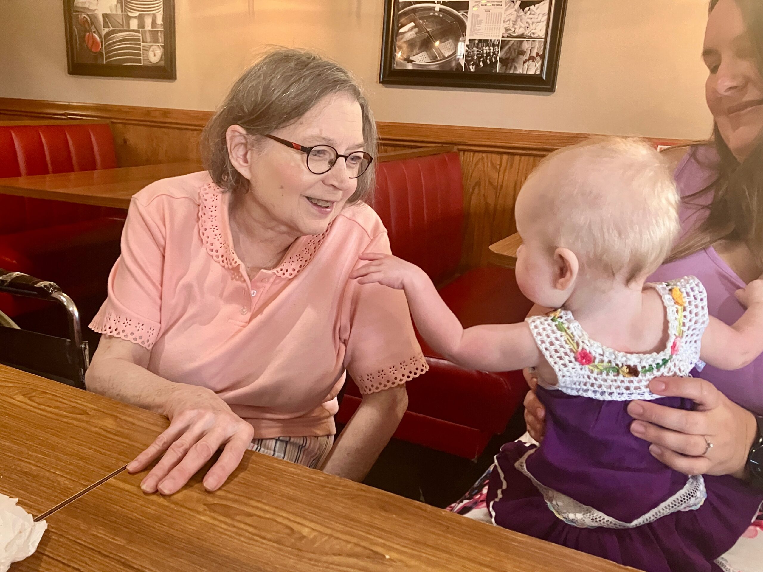 Older woman with short gray hair and glasses smiles at a child in a purple dress as they sit at a wooden restaurant table in a booth.
