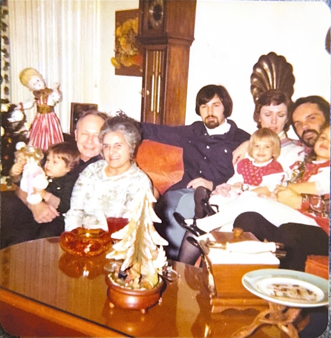 A multi‑generational family sits on a couch in a cozy living room during Christmas, with a small decorative tree on the coffee table in front of them.