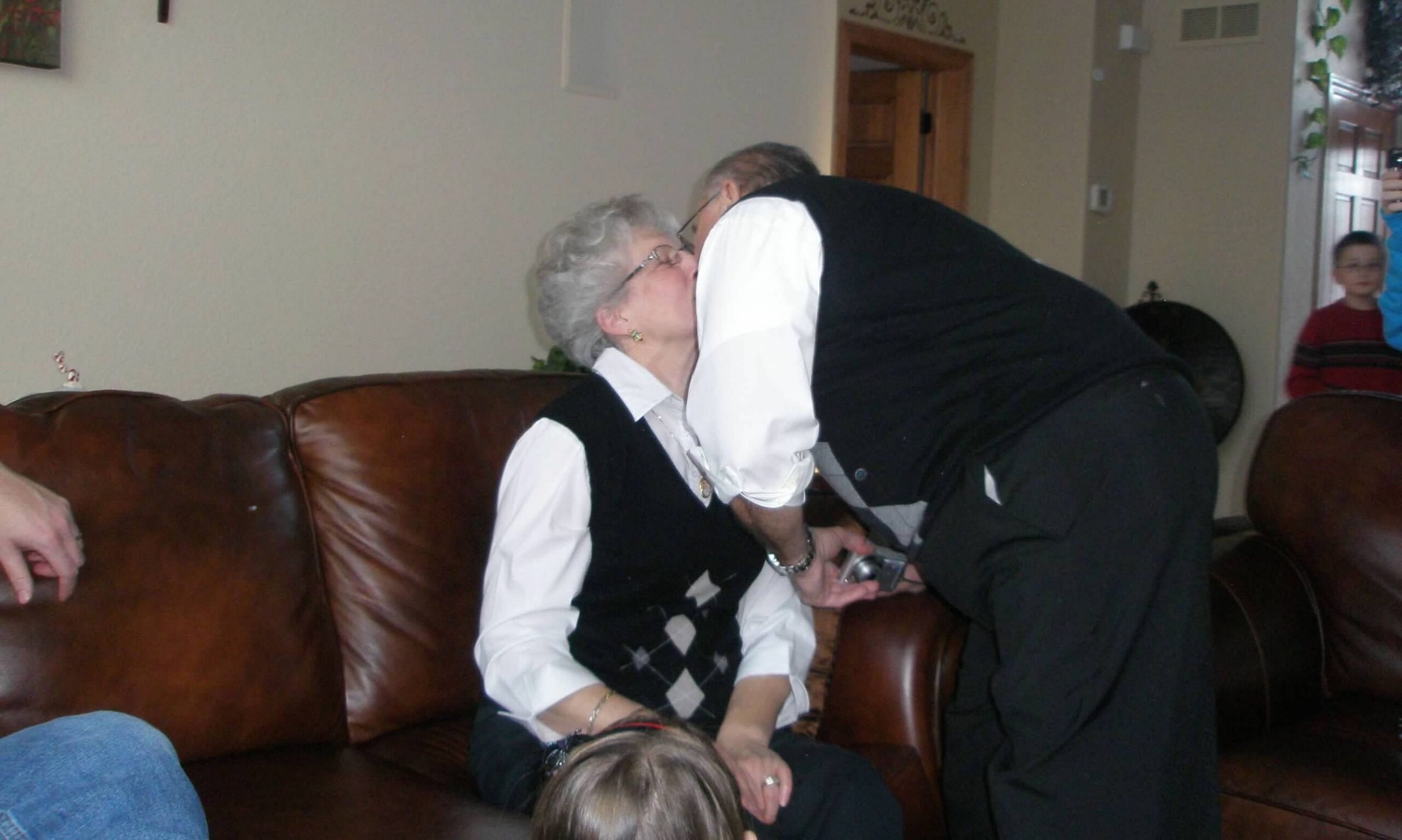 Two elderly people share a kiss on a brown leather couch in a living room, while a child stands in the doorway watching.