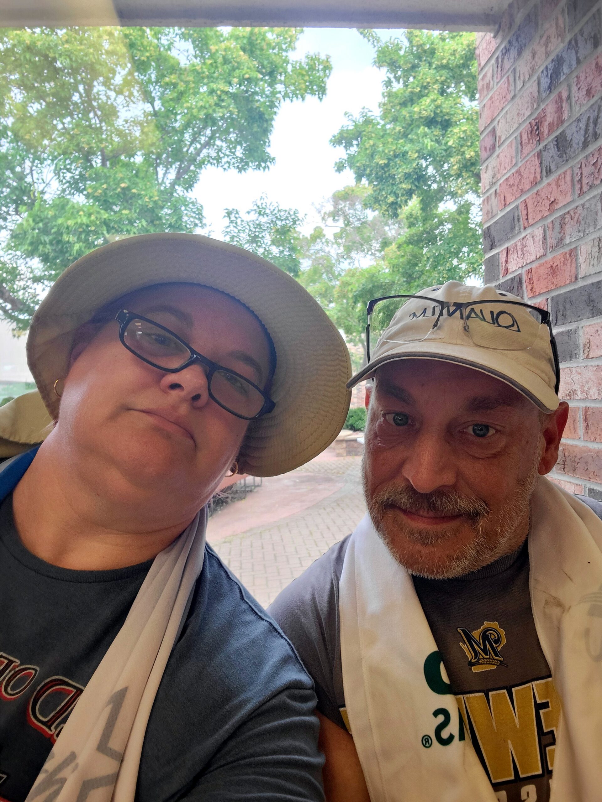 Close-up selfie of two people wearing hats, smiling, with green trees and a brick wall in the background.