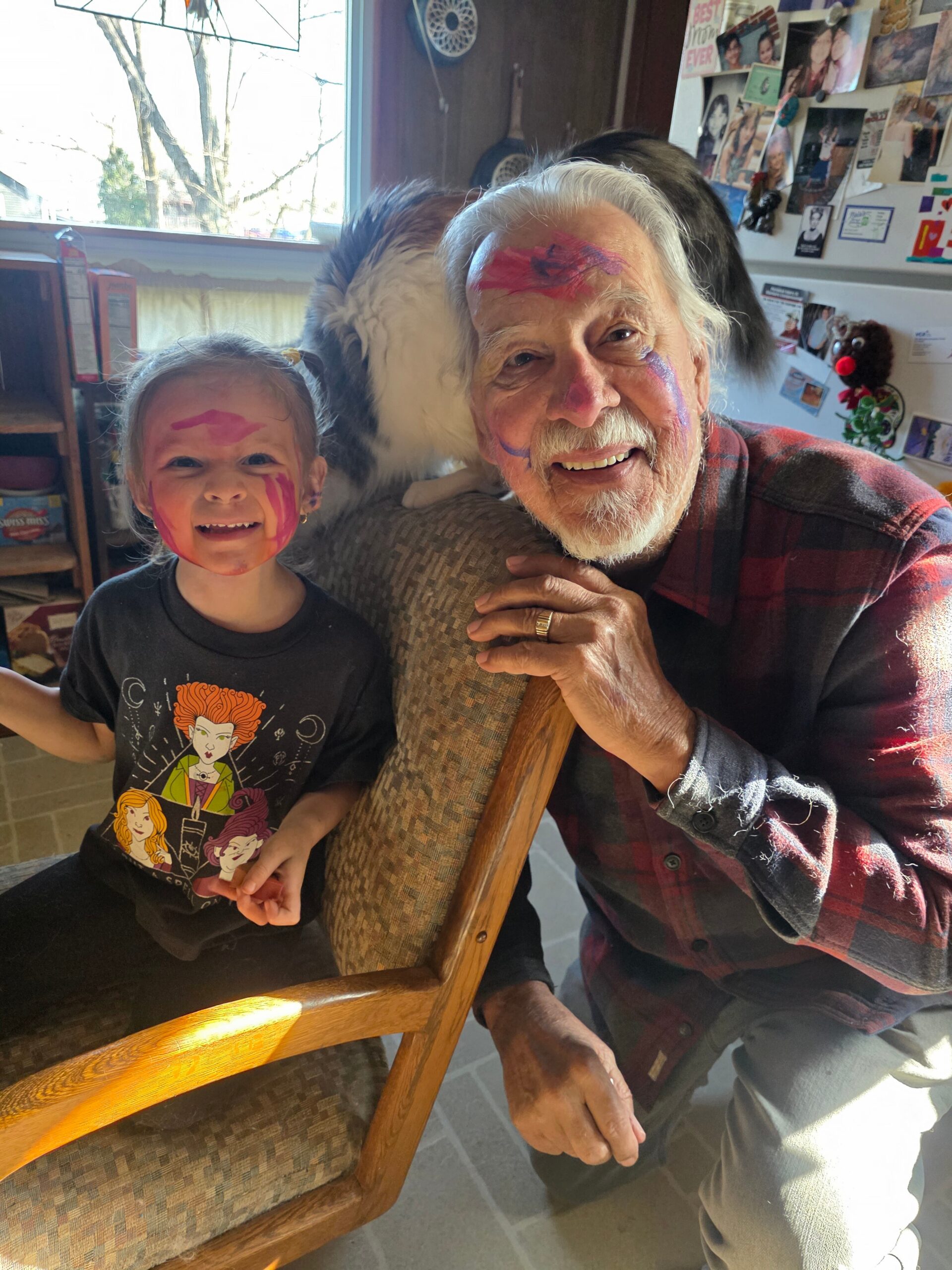Smiling young girl with bright pink face paint beside an elderly man with painted forehead and cheeks, posing indoors near a sofa.