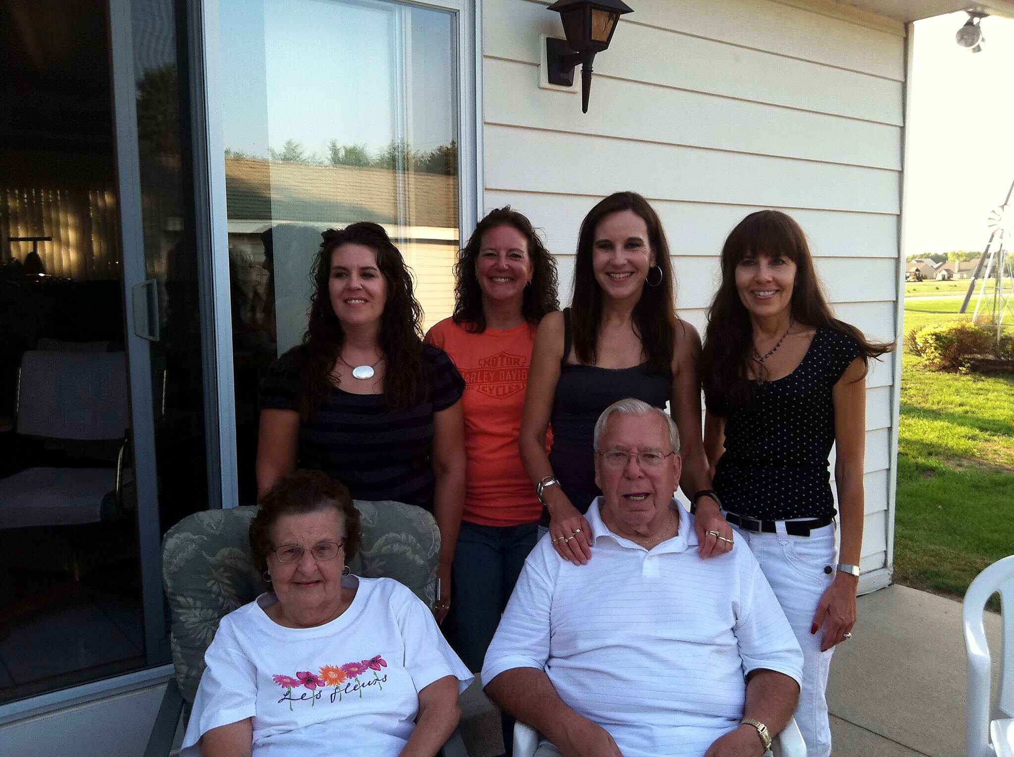 Group of adults posing on a house’s porch outdoors, with a sliding door behind and a lawn to the right.