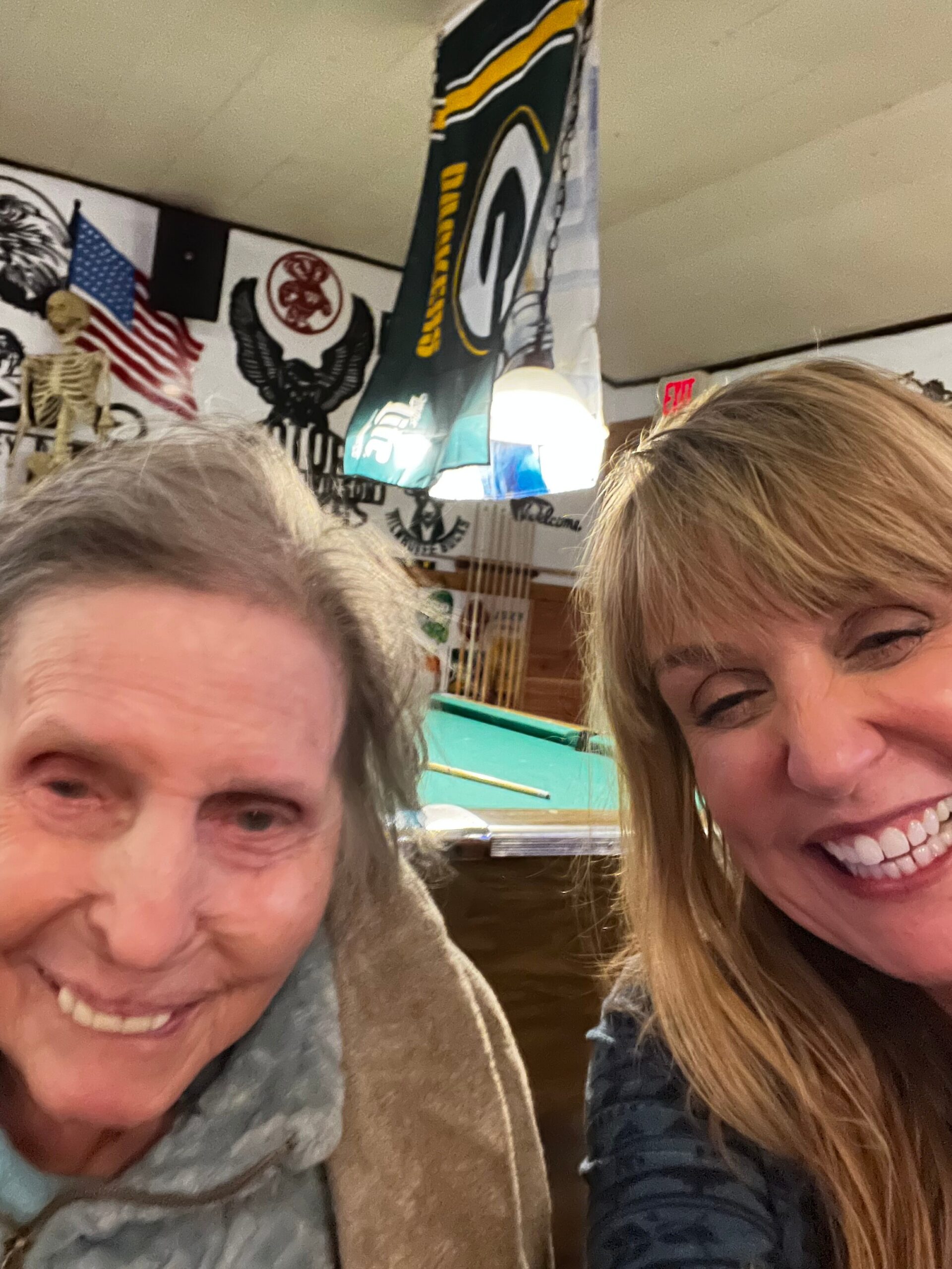 Two people smiling for a selfie in a room with a pool table, flags, and wall art in the background.